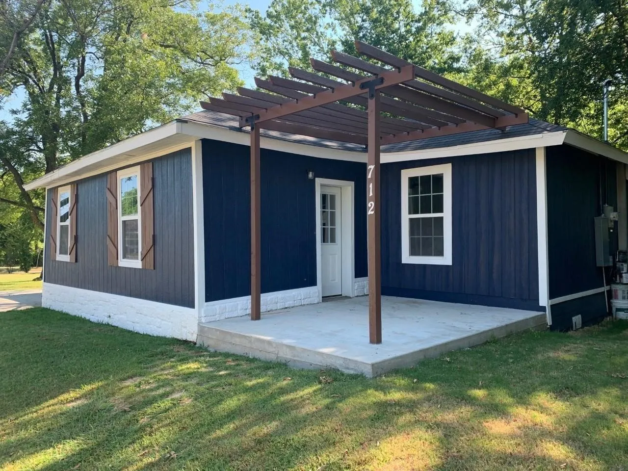 Blue vertical siding with white trim, concrete patio, wood pergola, and decorative window shutters