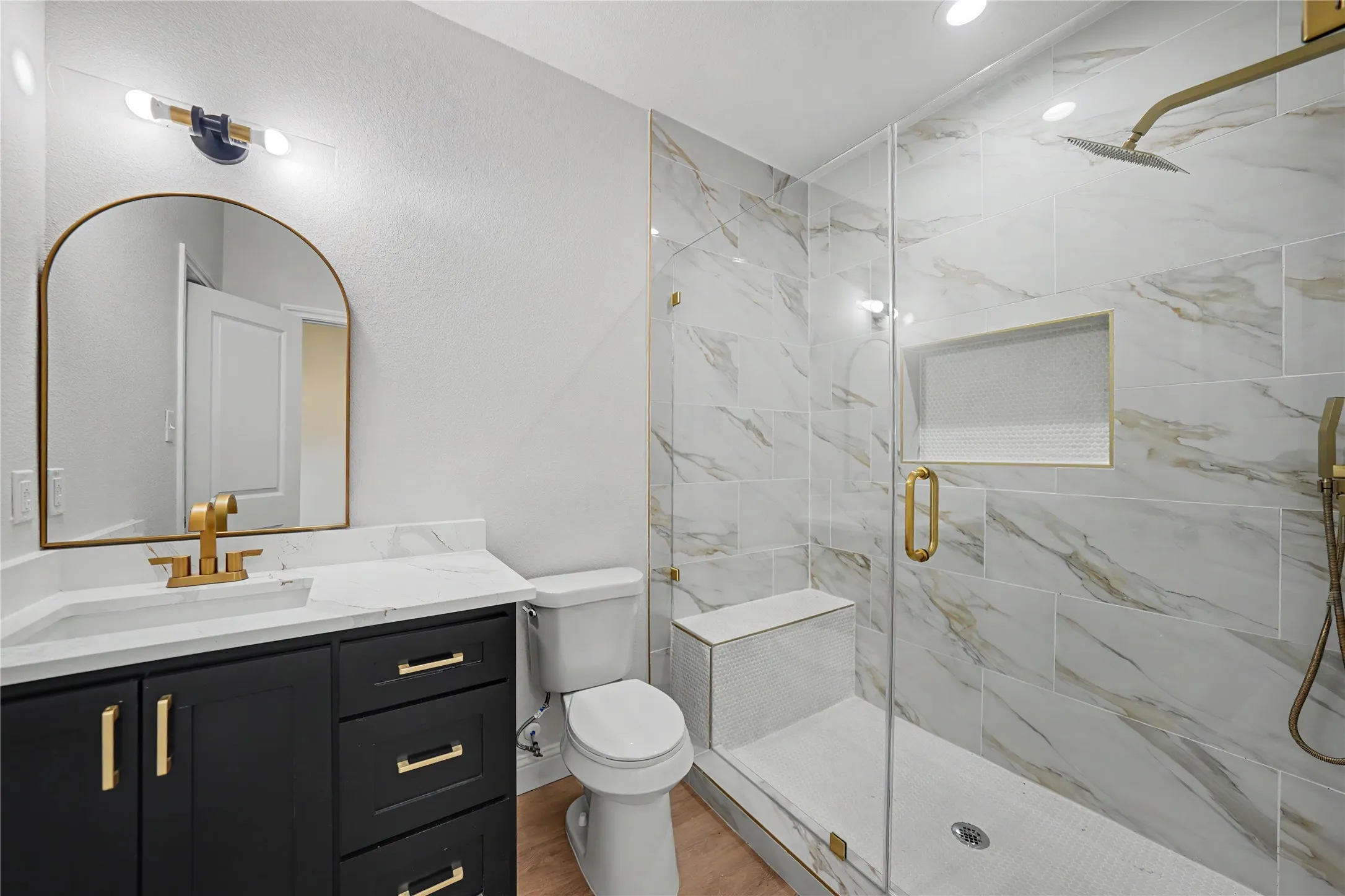 Modern bathroom featuring a black vanity with gold hardware, a white countertop, an arched gold-framed mirror, and a glass-enclosed shower with white and gold-veined tile