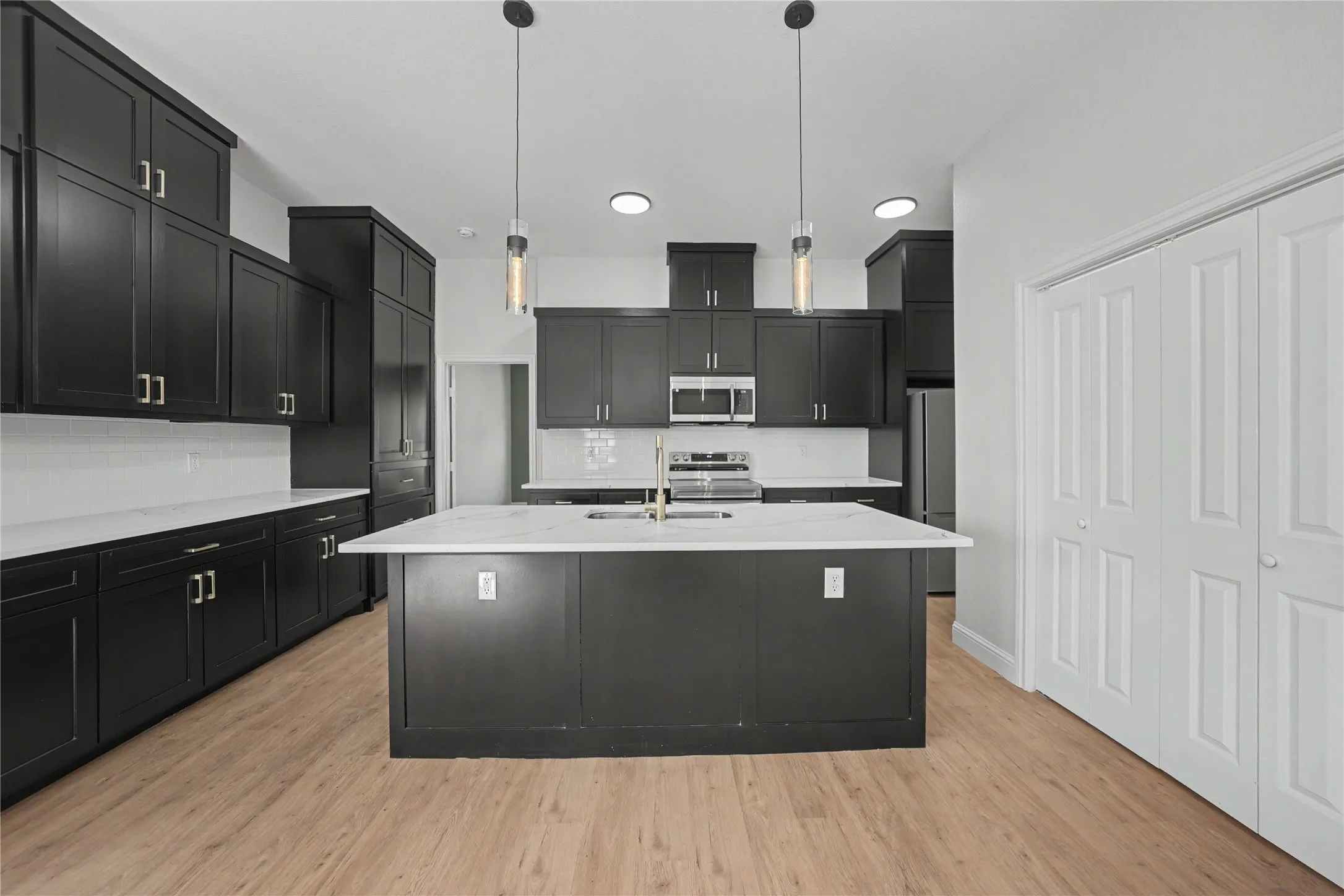 Modern kitchen featuring dark cabinetry, white countertops, stainless steel appliances, wood-finish flooring, and pendant lighting