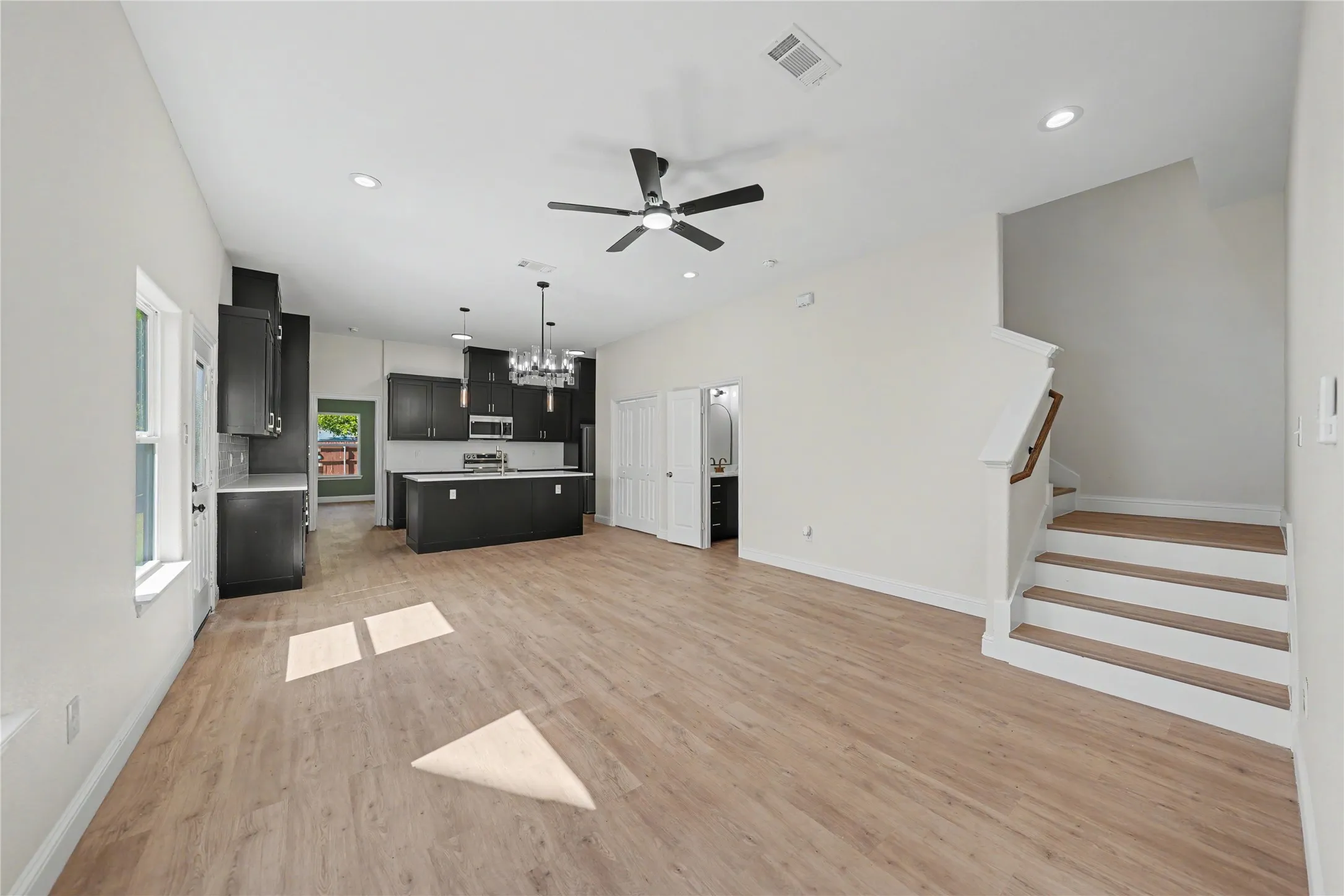 Open-concept living space featuring wood-finish flooring, a modern kitchen with dark cabinetry, stainless steel appliances, a kitchen island, and a staircase with a wood handrail