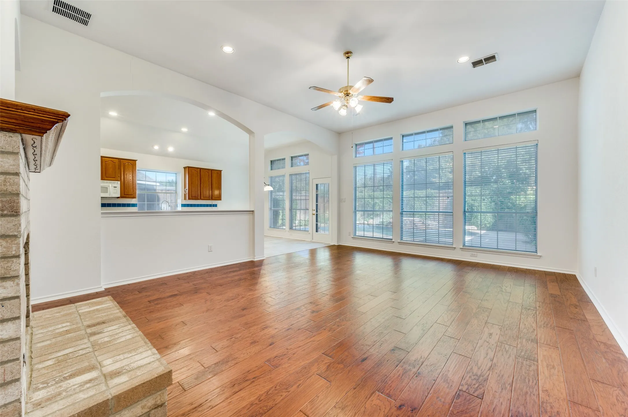 Varnished wood-finish flooring, vaulted ceiling with a ceiling fan, stone-clad fireplace, large windows with horizontal blinds, and recessed lighting