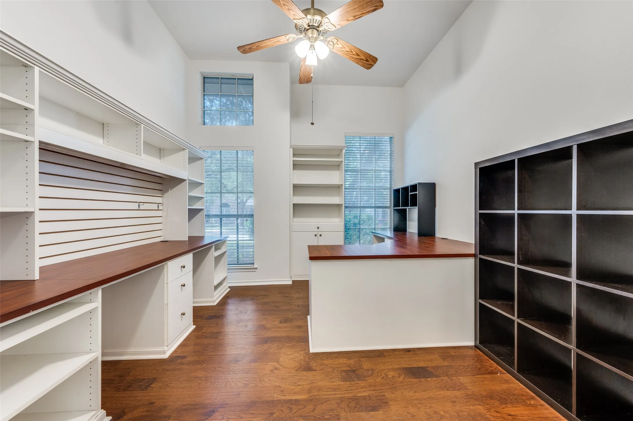 Dedicated workspace featuring extensive built-in shelving, two integrated desk surfaces, high ceilings, and wood-finish flooring
