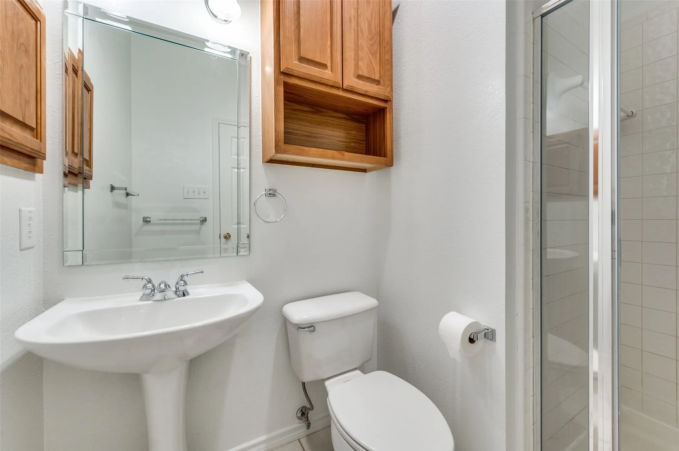 Pedestal sink with chrome faucet, mirrored vanity, wood-finish wall cabinetry, and a shower enclosure with white tile