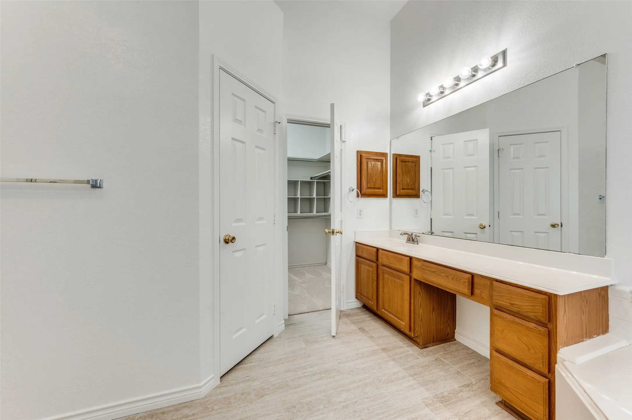 White paneled door, wood-finish vanity with ample storage, integrated sink, and a large wall-mounted mirror with overhead lighting.  Nice large walk-in closet through the open door.