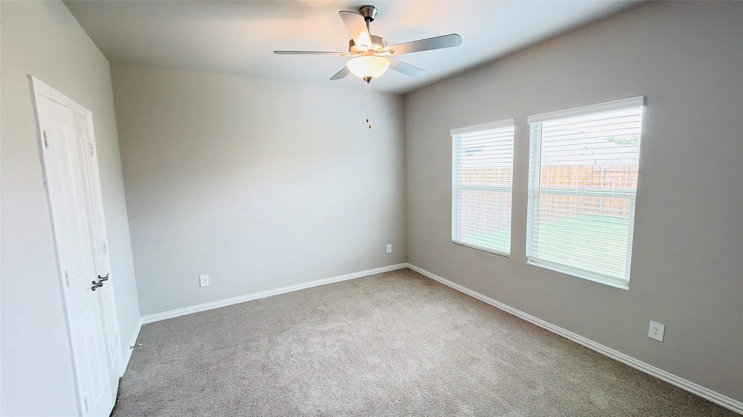 Spacious room with neutral wall tones, carpeted flooring, two windows with blinds, and a ceiling fan with integrated lighting