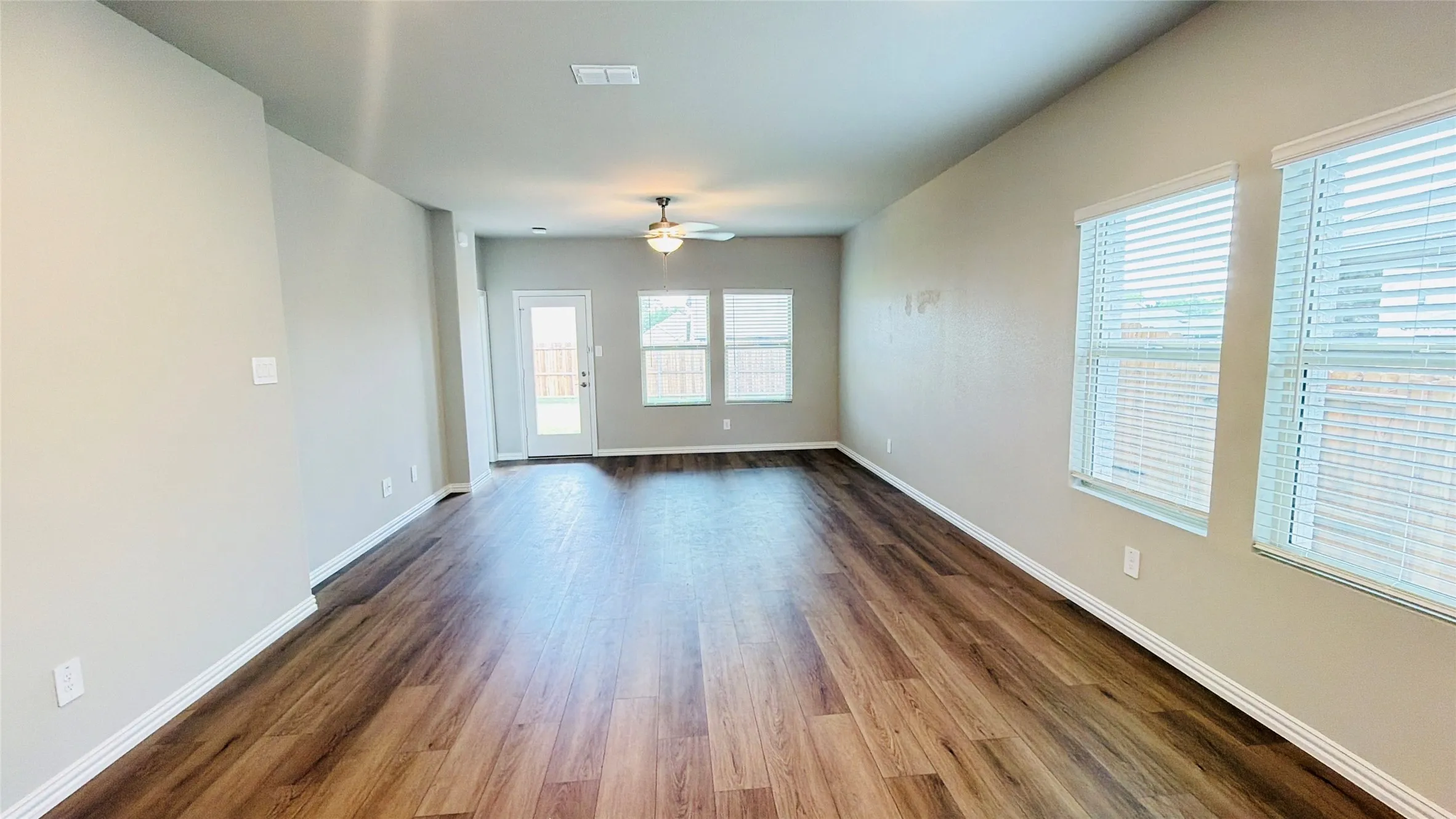 Spacious room featuring wood-finish flooring, light neutral walls, multiple windows with blinds, a ceiling fan, and a glass-paneled exterior door