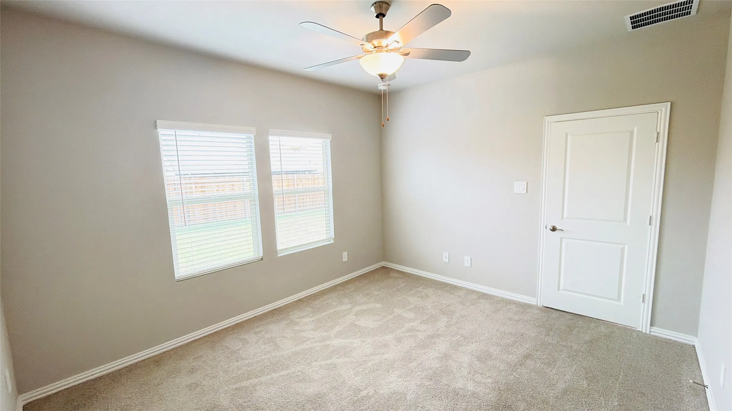 Neutral tone wall paint, carpet flooring, two windows with blinds, ceiling fan with integrated lighting, and a white interior door