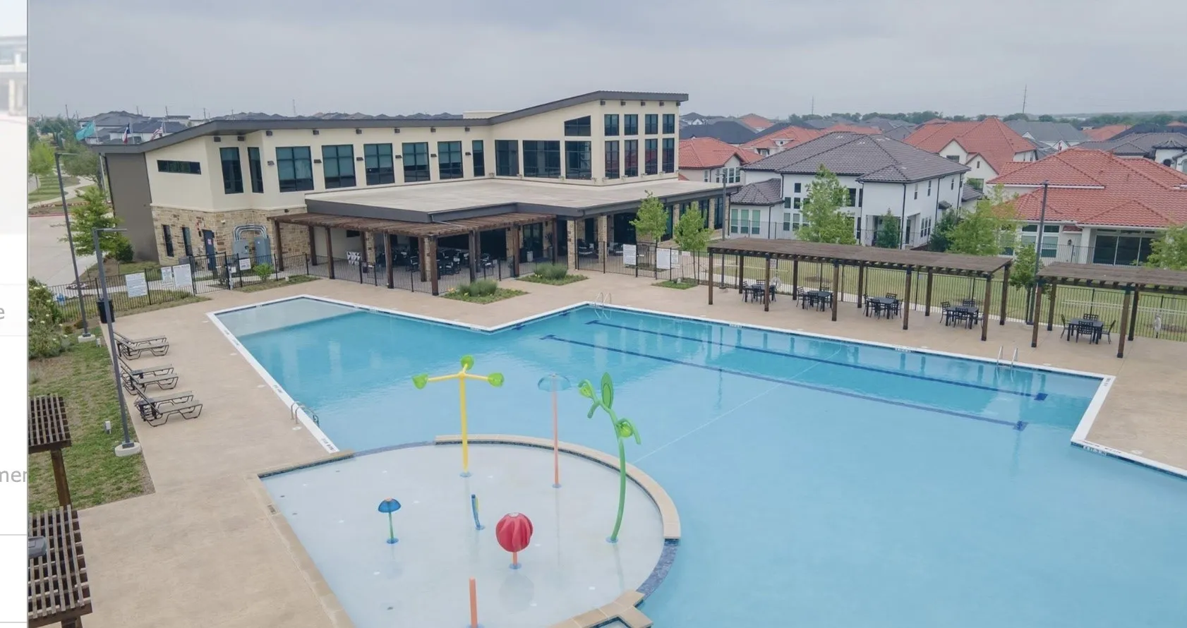 Expansive community pool with a dedicated splash pad, adjacent to a modern clubhouse featuring a covered patio with pergolas