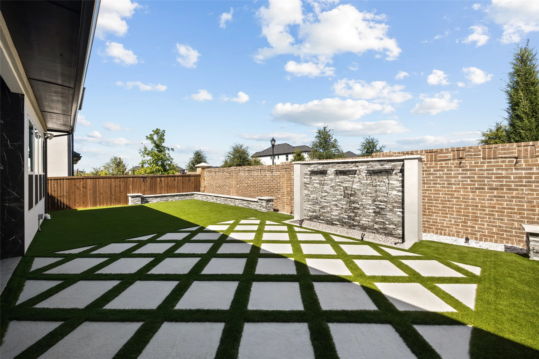 Expansive backyard featuring a water feature with stone-finish cladding, a brick perimeter wall, and a geometric pattern of concrete pavers set in artificial turf