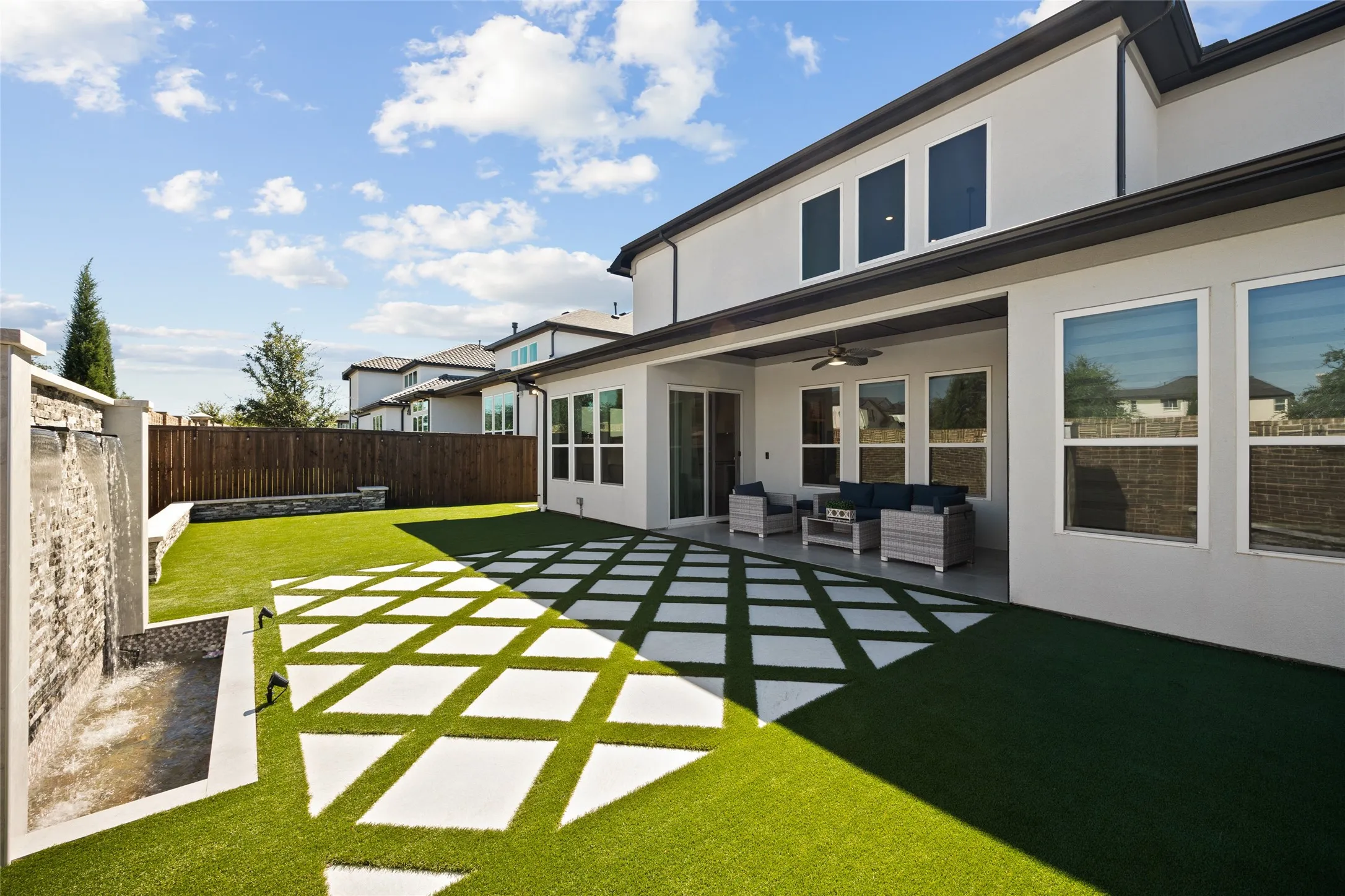 Private backyard featuring a stone-clad water feature, artificial turf, modern patio pavers, and a covered patio with an outdoor ceiling fan