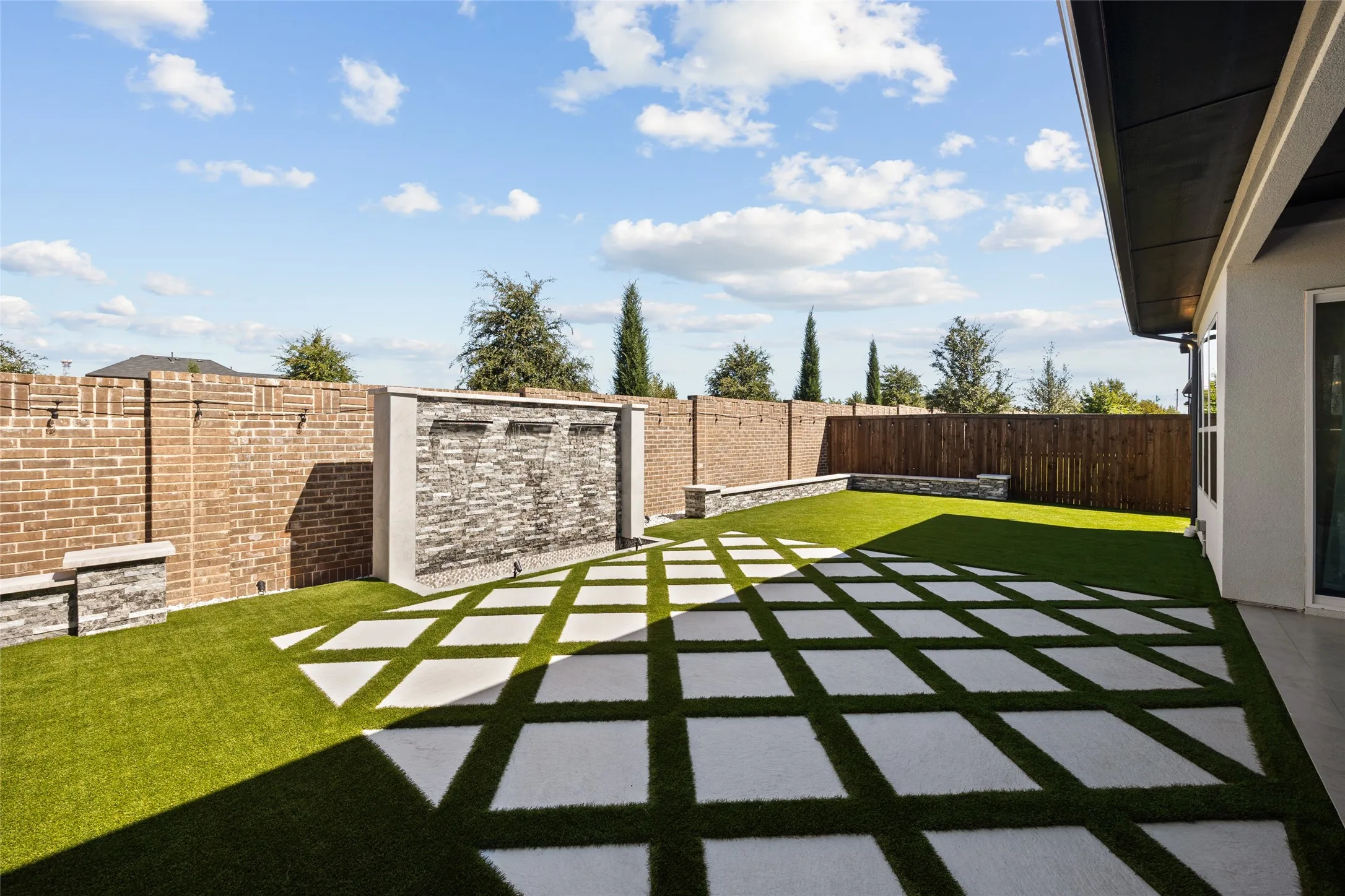 Expansive backyard featuring a brick perimeter wall, stacked stone water feature, artificial turf, and geometric paver patio