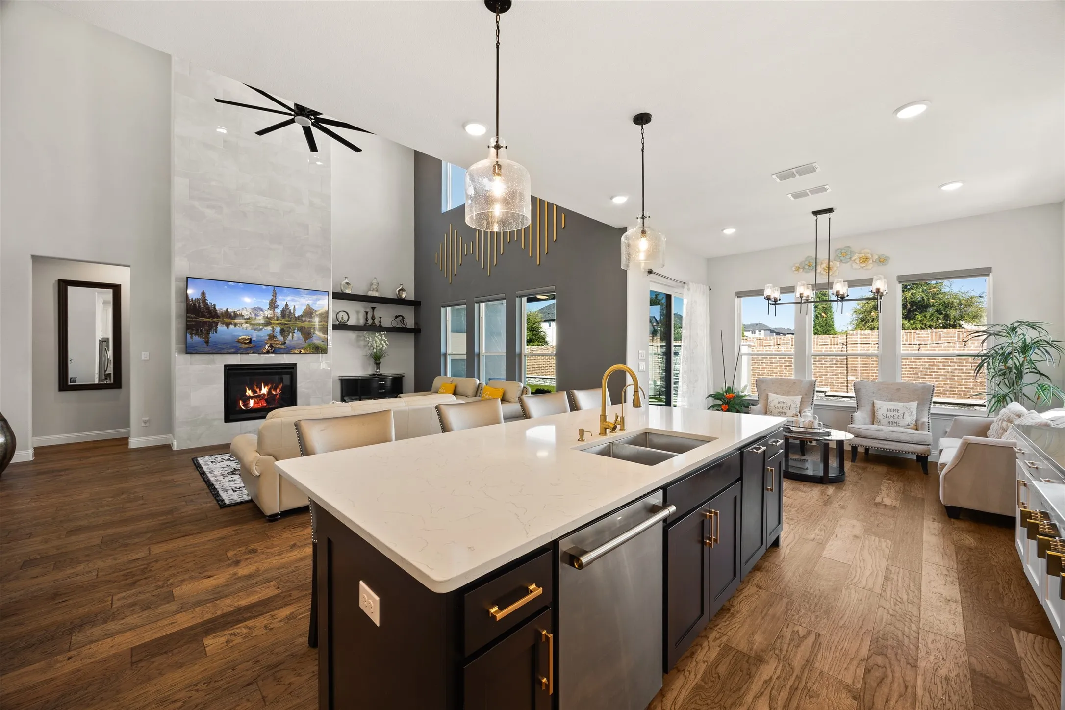 Expansive open-concept living area featuring high ceilings, wood-finish flooring, a contemporary fireplace with a full-height surround, and an island kitchen with a dishwasher and under-mount sink