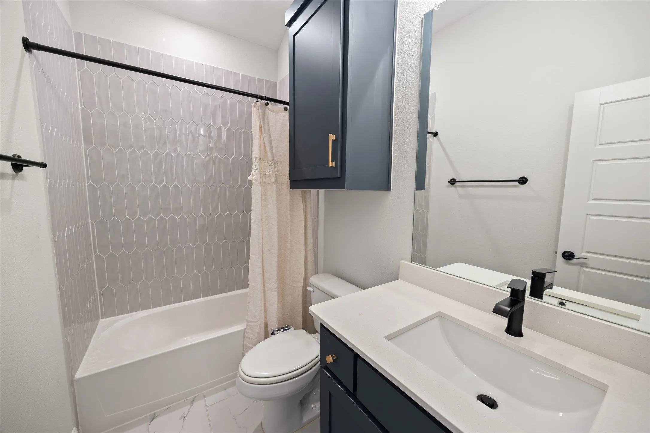 White bathroom featuring a single vanity with a white countertop and dark cabinet, a built-in tub with light grey elongated hexagonal tile surround, and a wall-mounted mirror