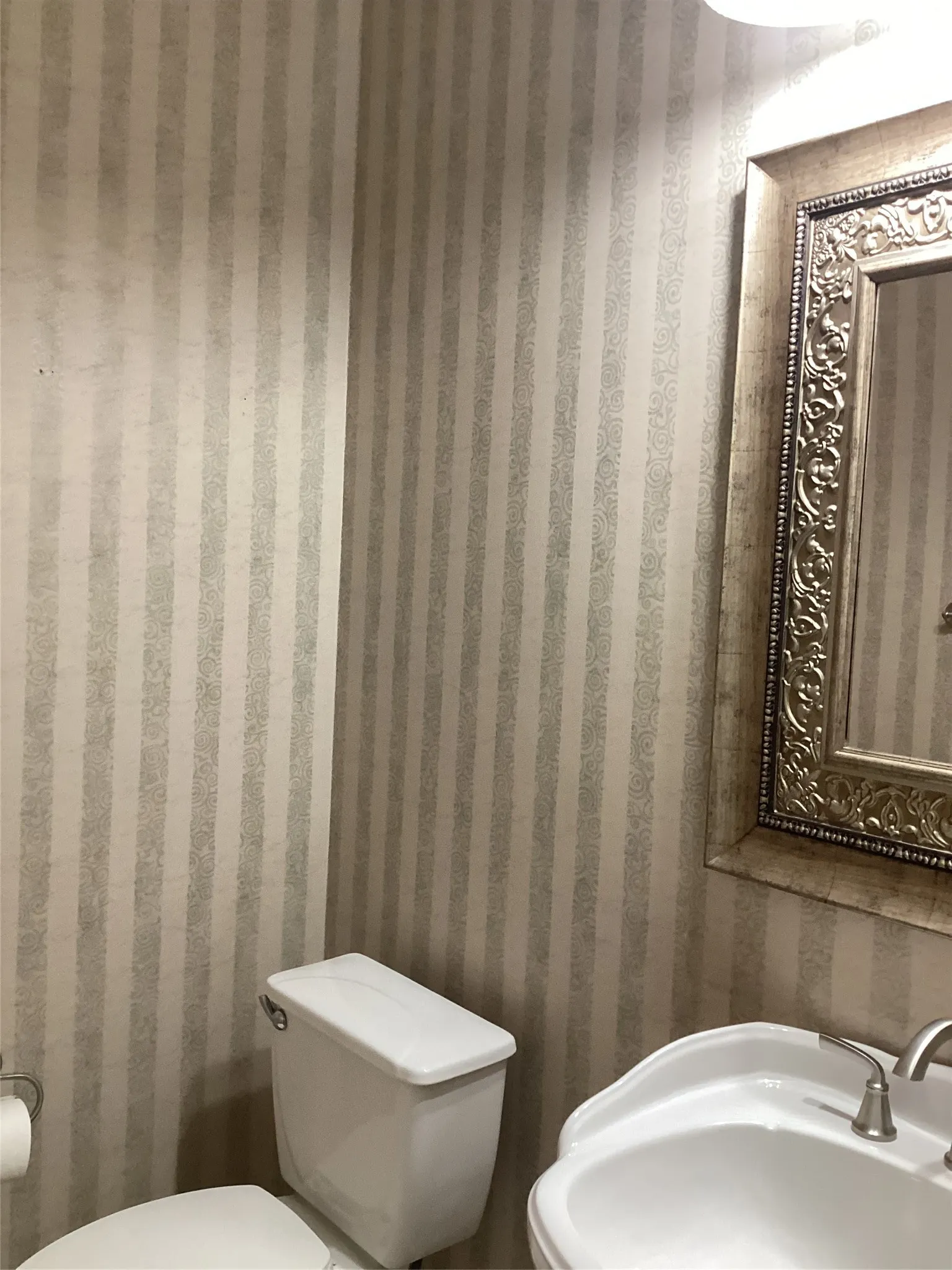 Pedestal sink with a brushed nickel faucet, ornate framed mirror, and vertical striped wallpaper