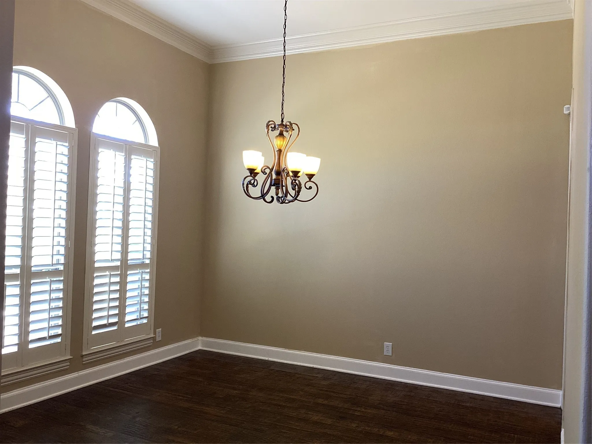 Wood-finish flooring, architectural arched windows with plantation shutters, crown molding, and a decorative chandelier