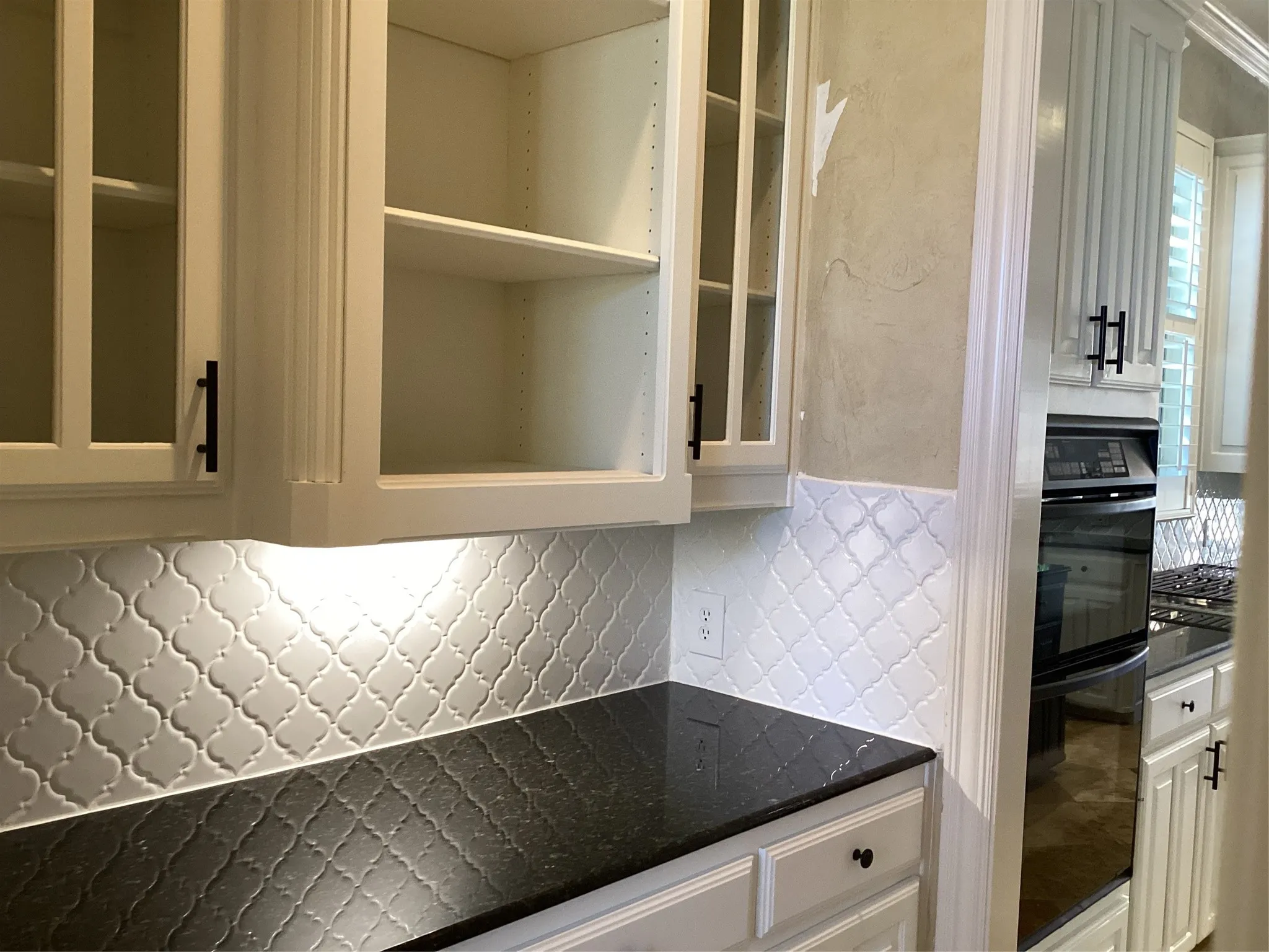 White cabinetry with glass-front uppers, dark countertops, white arabesque tile backsplash, and under-cabinet lighting