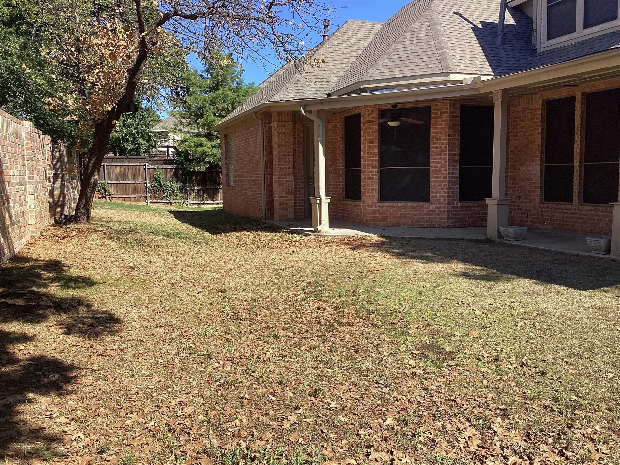 Expansive backyard featuring a brick privacy fence, mature trees, and a covered patio with a ceiling fan