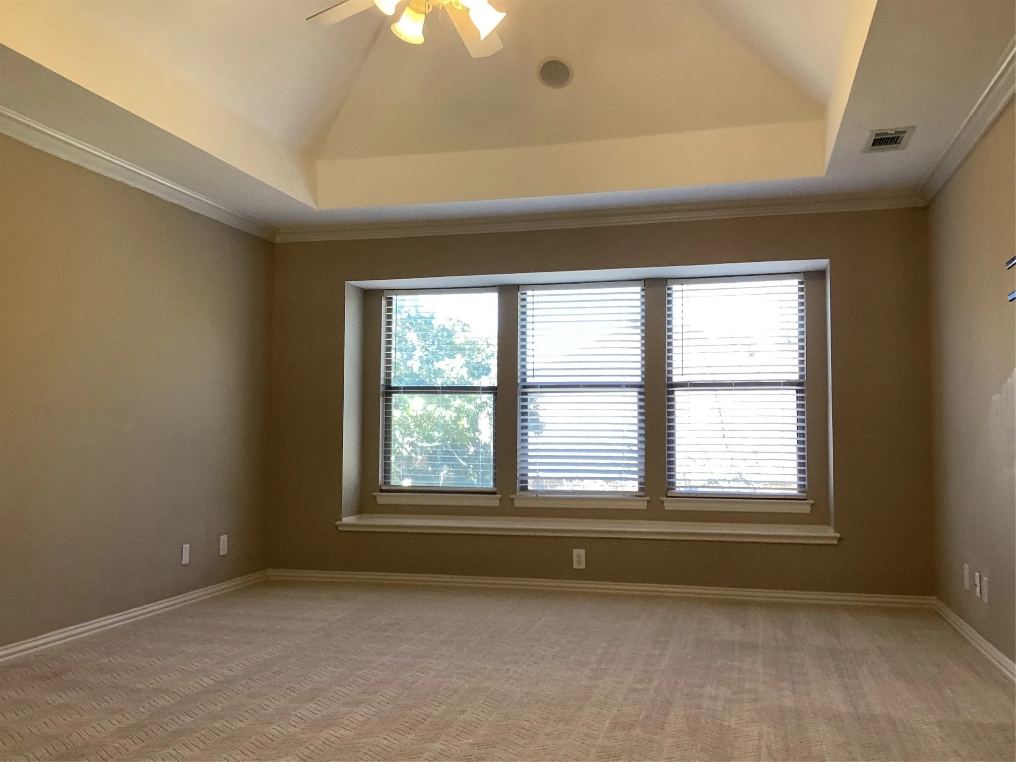 Tray ceiling with crown molding, ceiling fan with integrated lighting, triple window with blinds, window seat, and neutral-toned carpeting