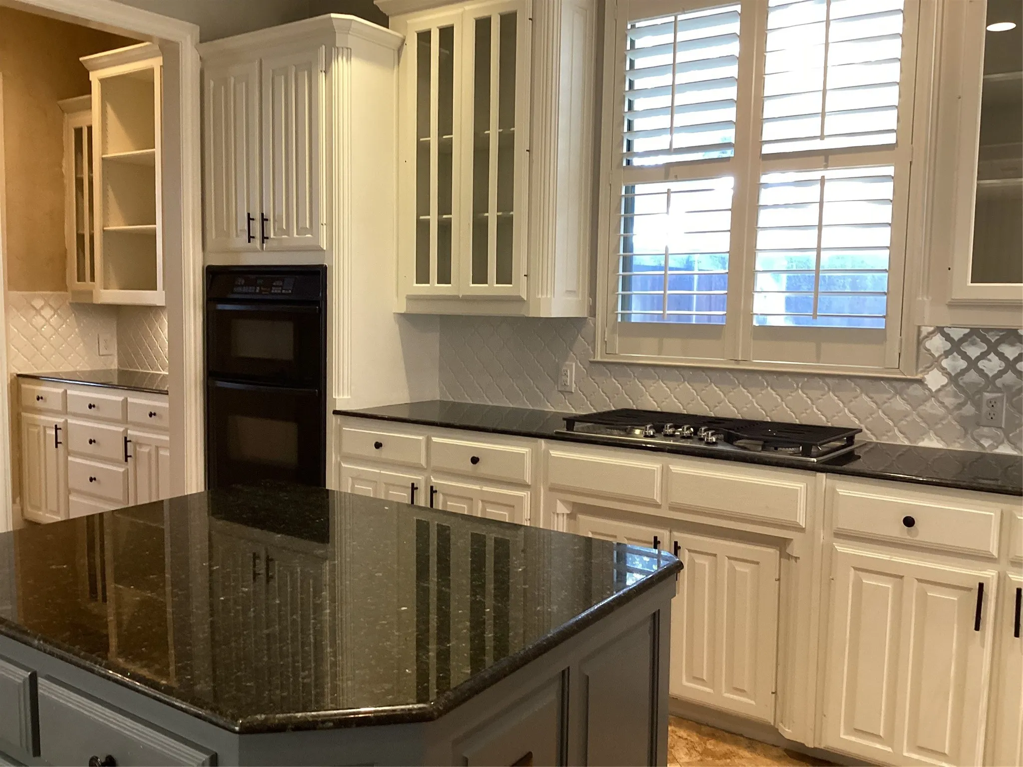 White cabinetry with varied door styles, black double wall ovens, island with dark stone countertop, white arabesque tile backsplash, and plantation shutters