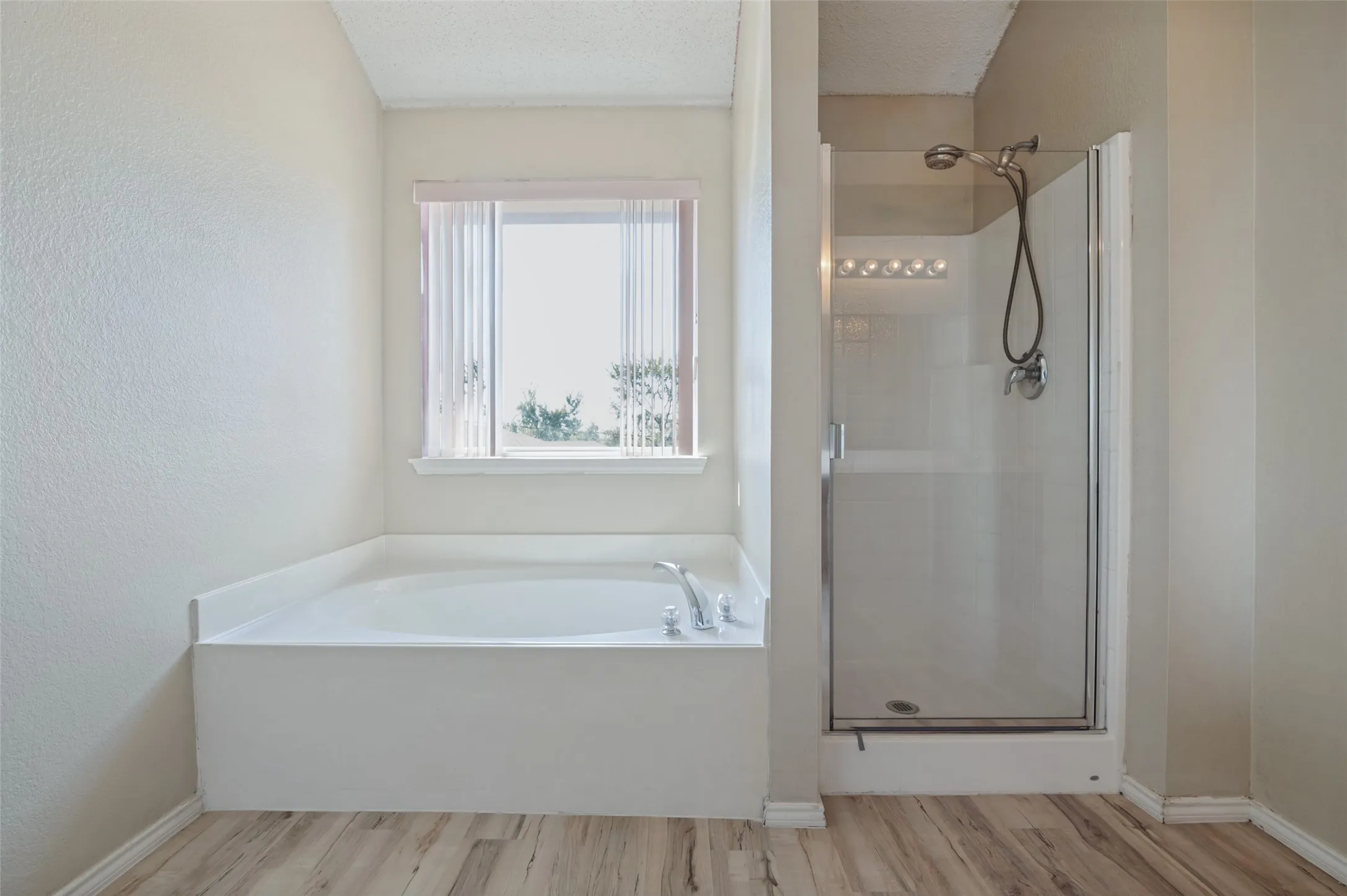 Soaking tub with window, separate glass-enclosed shower, wood-finish flooring, and wall-mounted light fixture