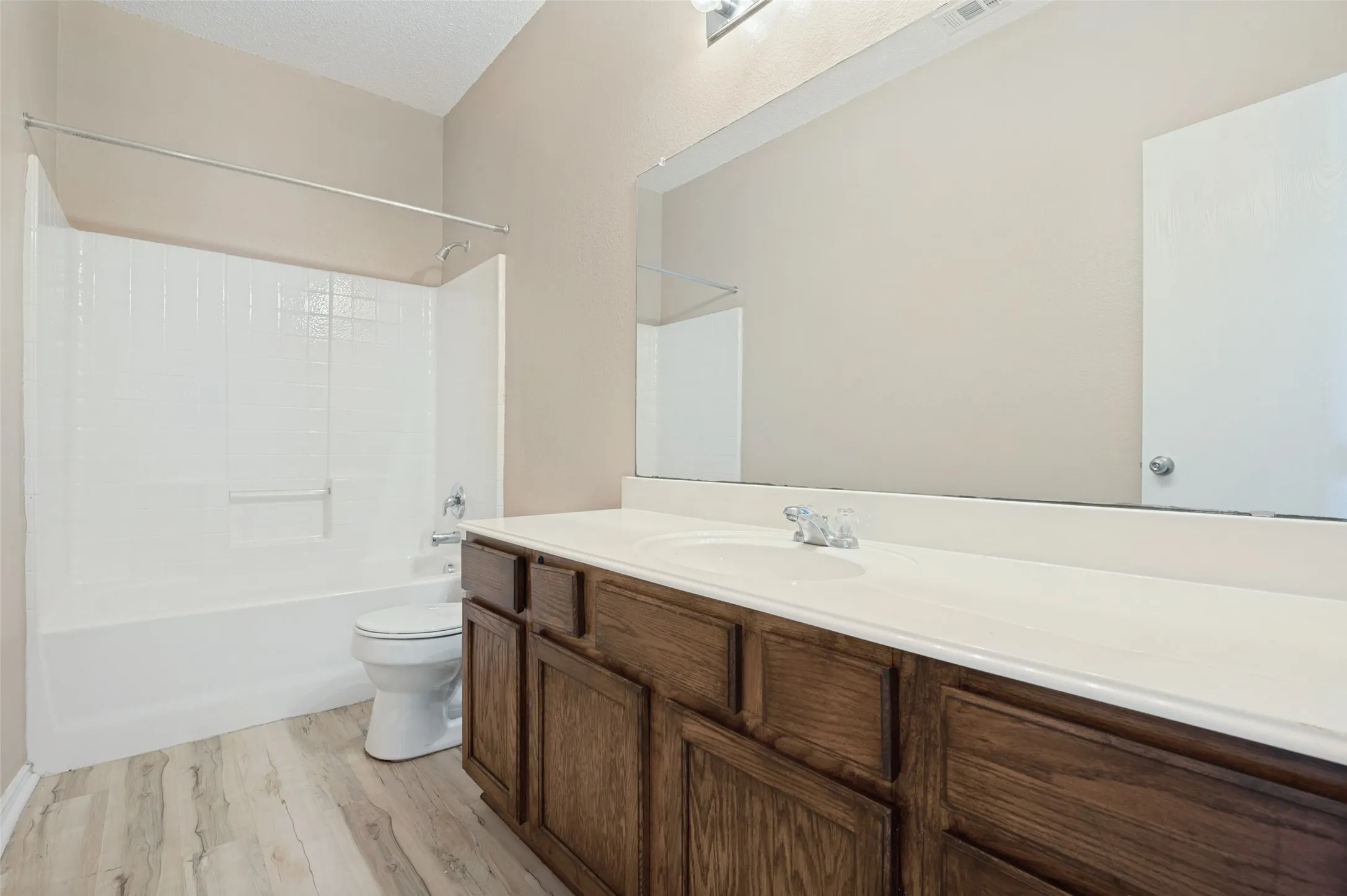 Wood-finish flooring, dark wood cabinetry, white countertop with integrated sink, full-width vanity mirror, and a white tub with shower