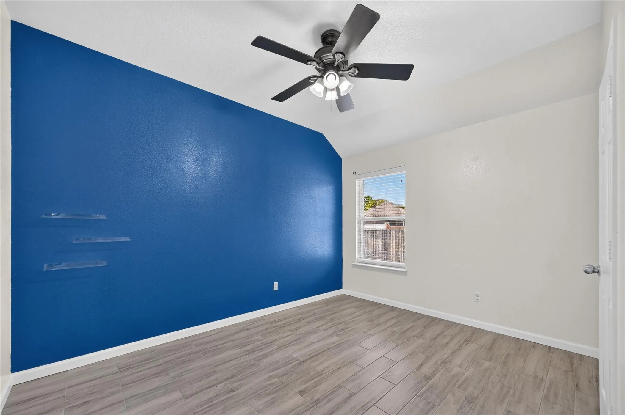 Vaulted ceiling, wood-finish tile flooring, ceiling fan with integrated lighting, window with blinds, and clear wall-mounted shelving