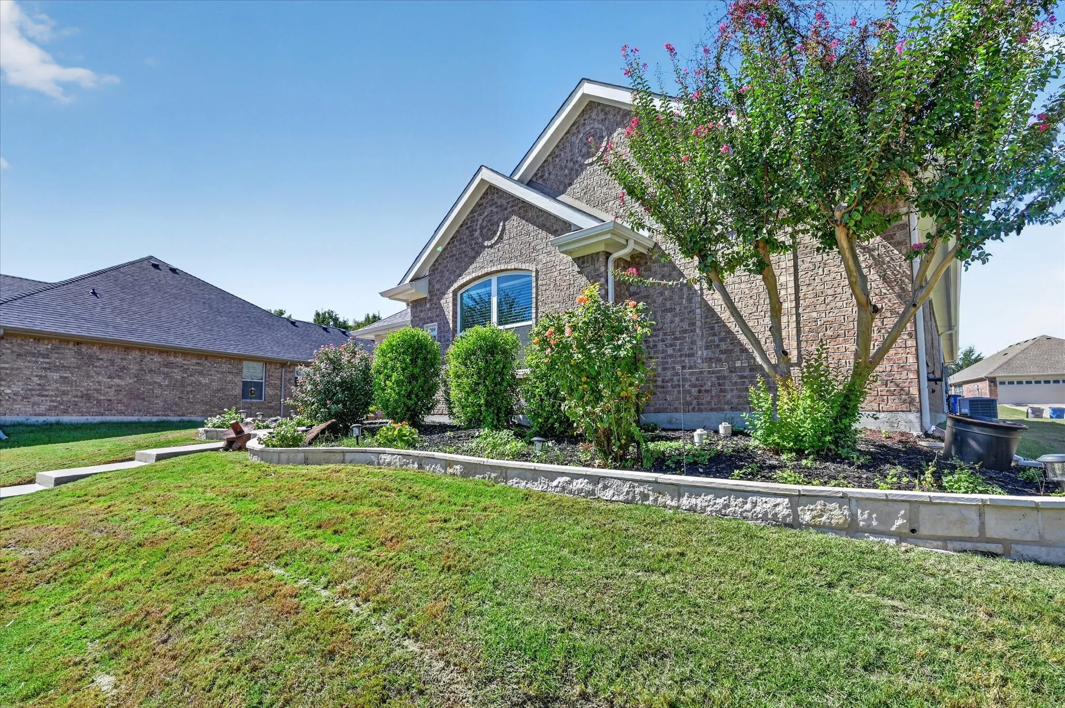 Brick exterior with arched window, concrete walkway, and landscaping featuring mature trees and shrubs