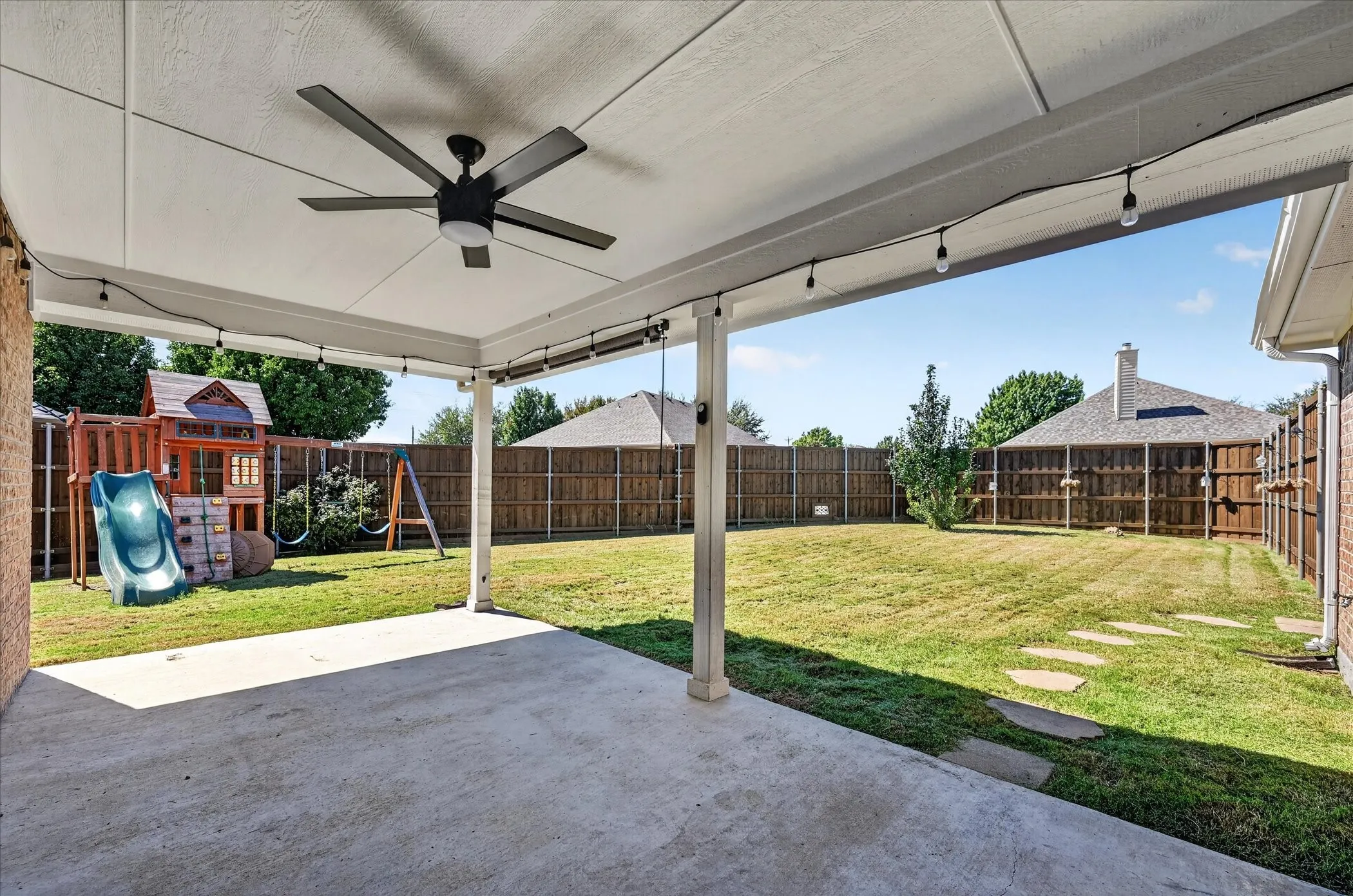 Expansive covered patio with ceiling fan and concrete flooring overlooking a large fenced yard with mature landscaping, wooden play structure, and swing set