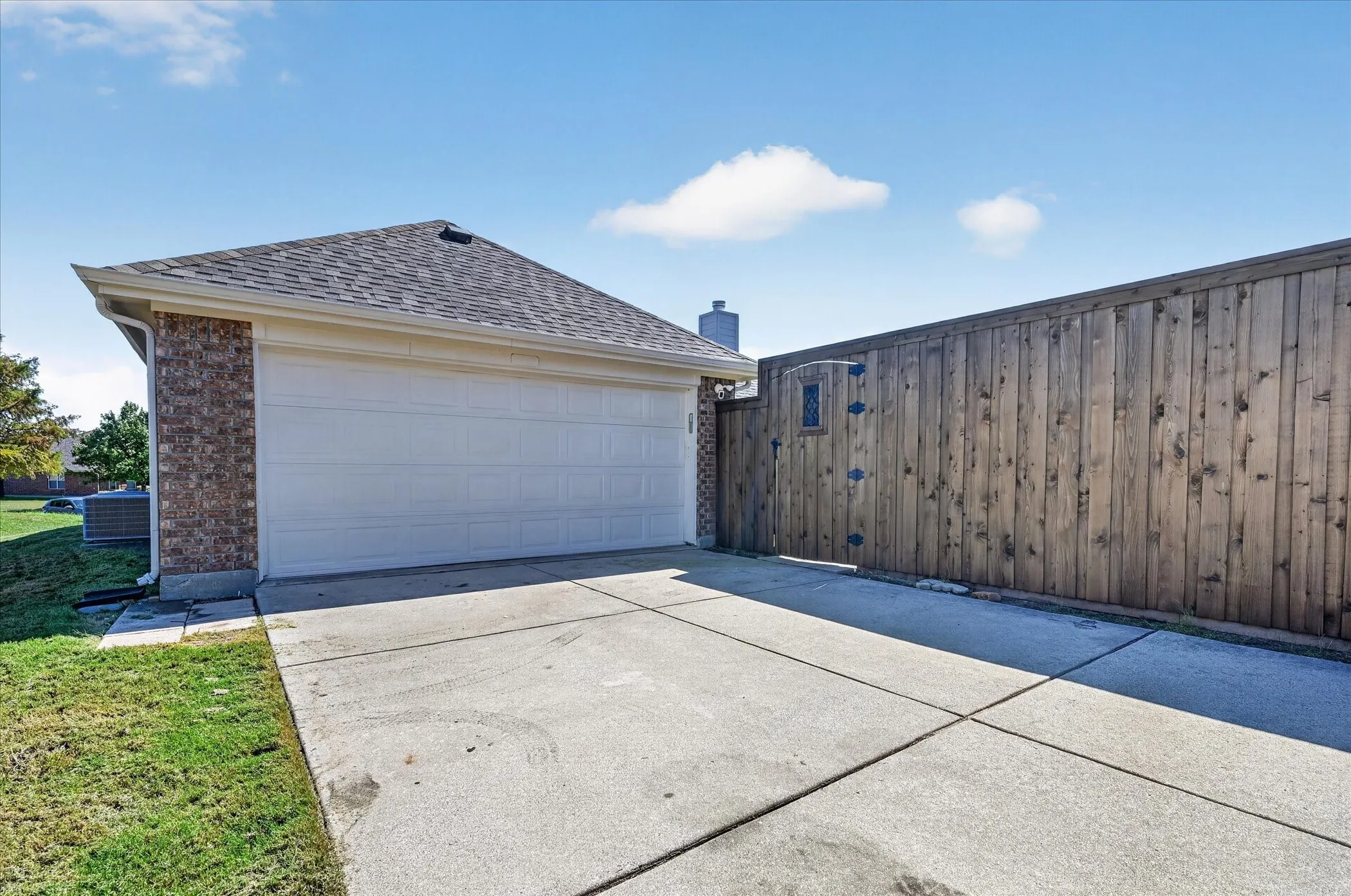 Attached two-car garage with a concrete driveway, brick exterior, and a wood privacy fence featuring an integrated decorative gate