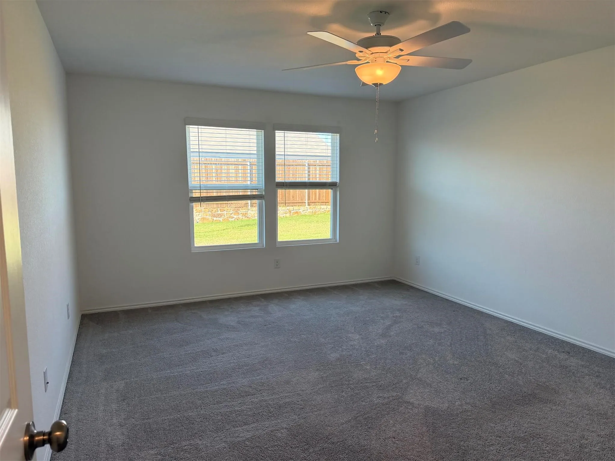 Spacious room featuring neutral carpeting, white walls, two windows with blinds, and a ceiling fan with integrated lighting