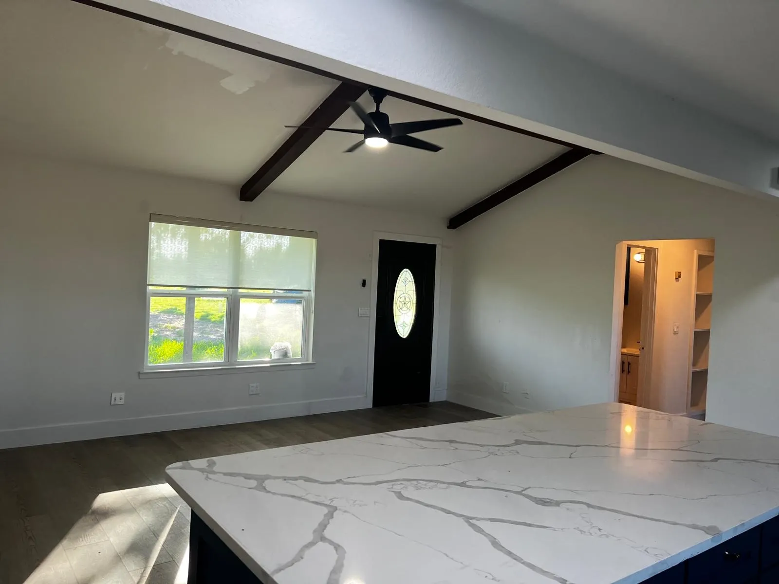 Vaulted ceiling with exposed beams and a ceiling fan, wood-finish flooring, white walls, and a front entry door with an oval glass insert