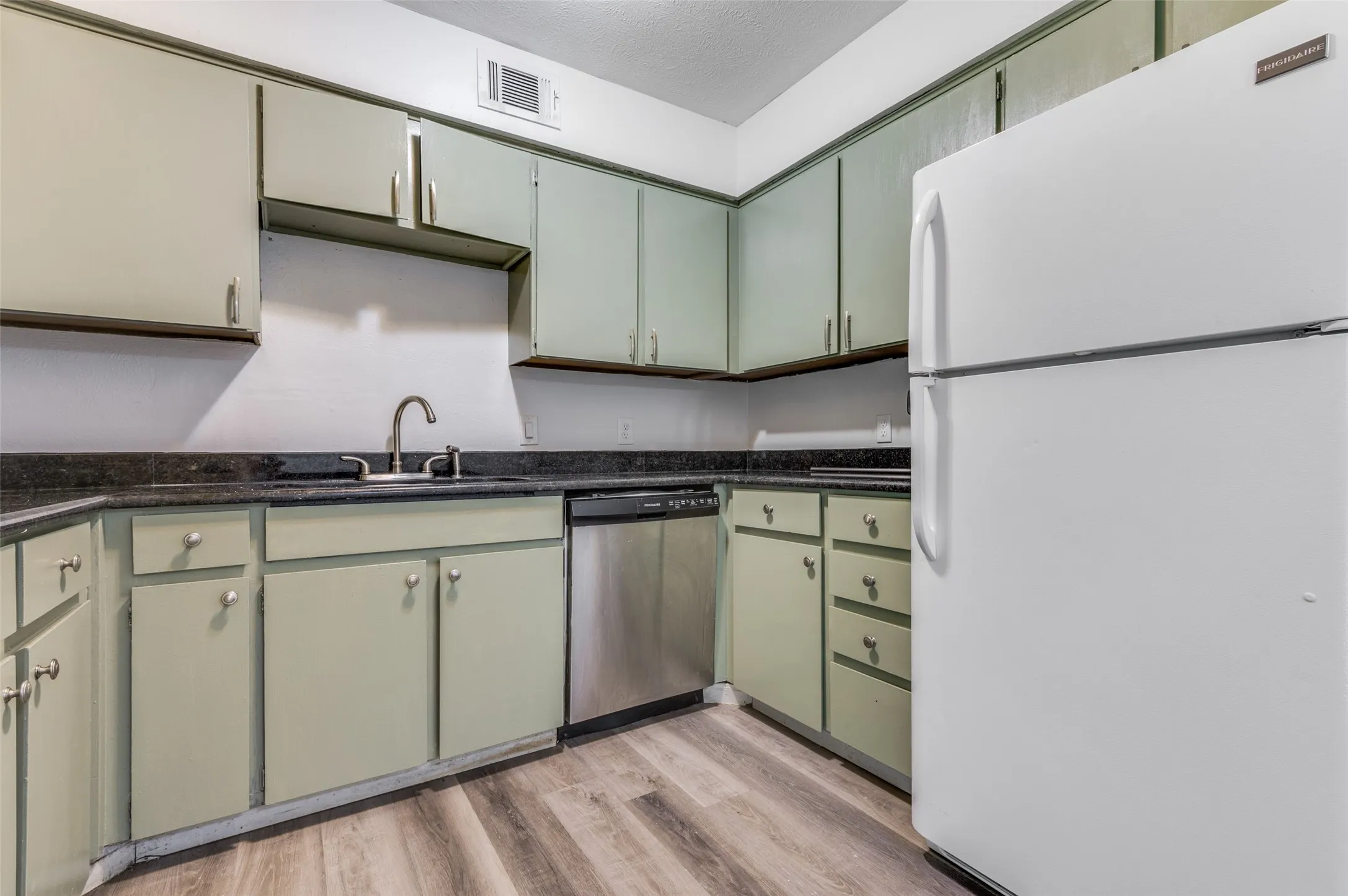 Kitchen featuring wood-finish flooring, light-toned cabinetry, dark countertops, a stainless steel dishwasher, and a white refrigerator