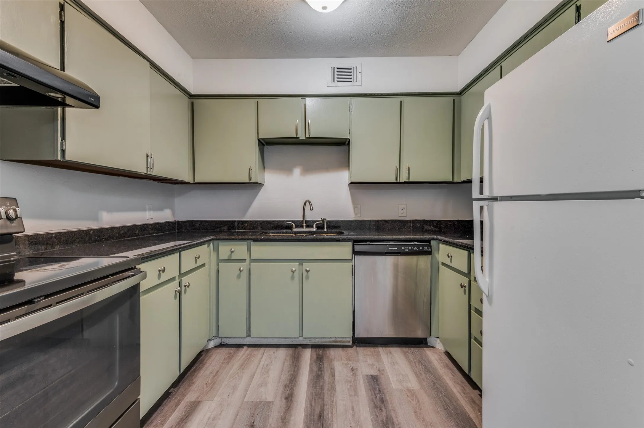 U-shaped kitchen featuring light green cabinetry, dark countertops, wood-finish flooring, and stainless steel dishwasher