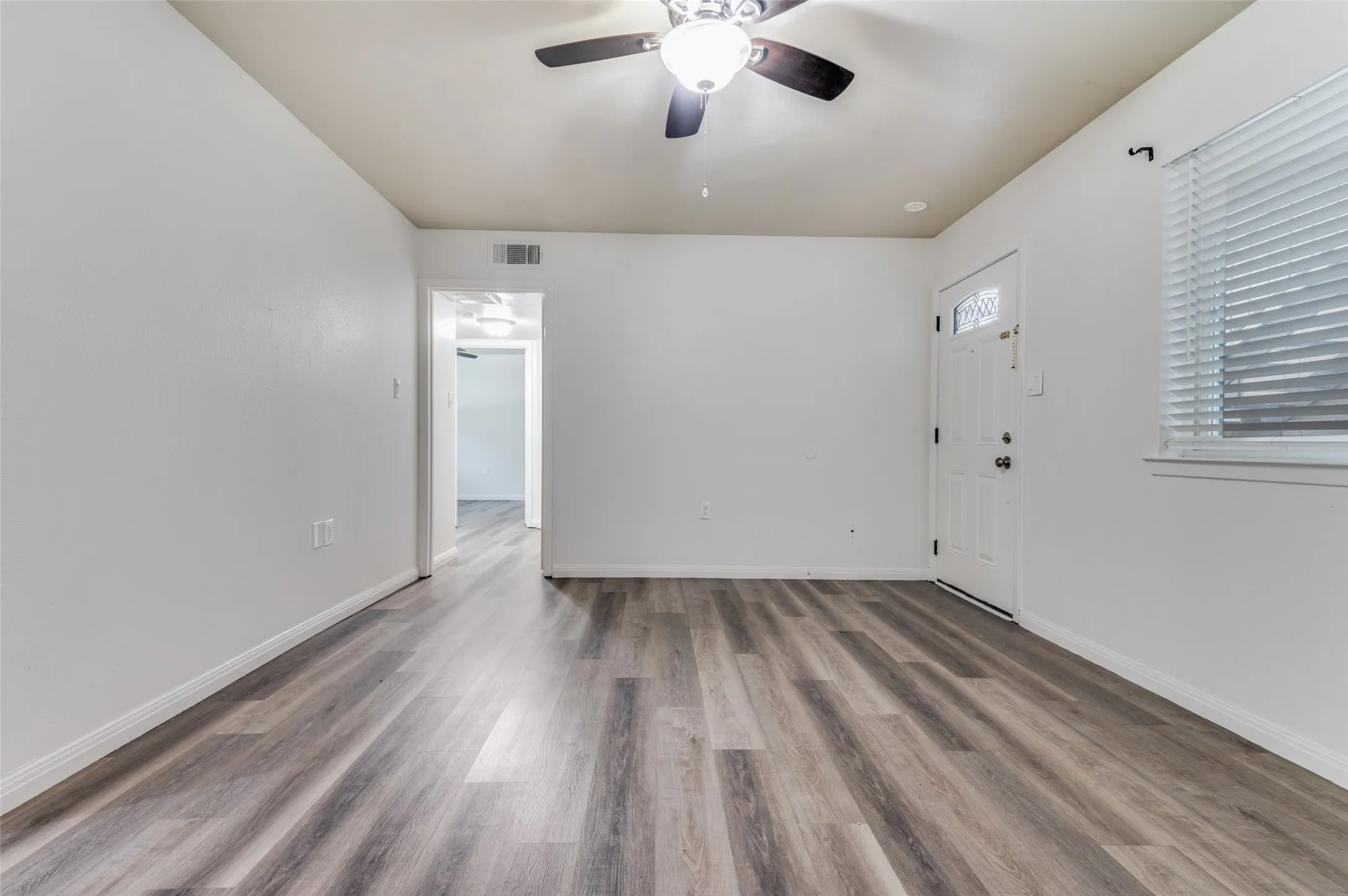 Wood-finish flooring, white walls, ceiling fan with integrated lighting, window with blinds, and a paneled entry door with decorative glass inserts