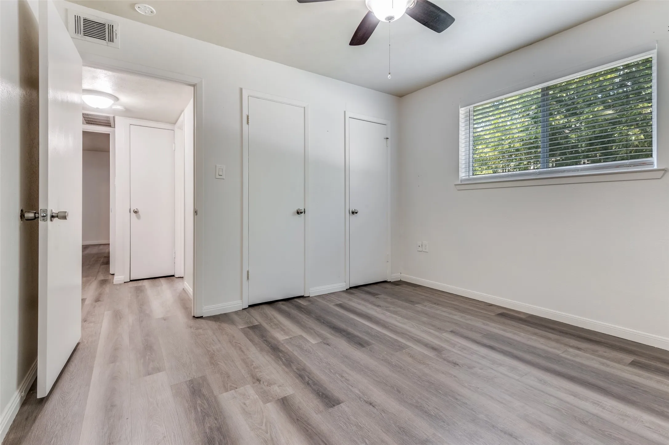 Wood-finish flooring, white painted walls, window with blinds, ceiling fan, and multiple interior doors