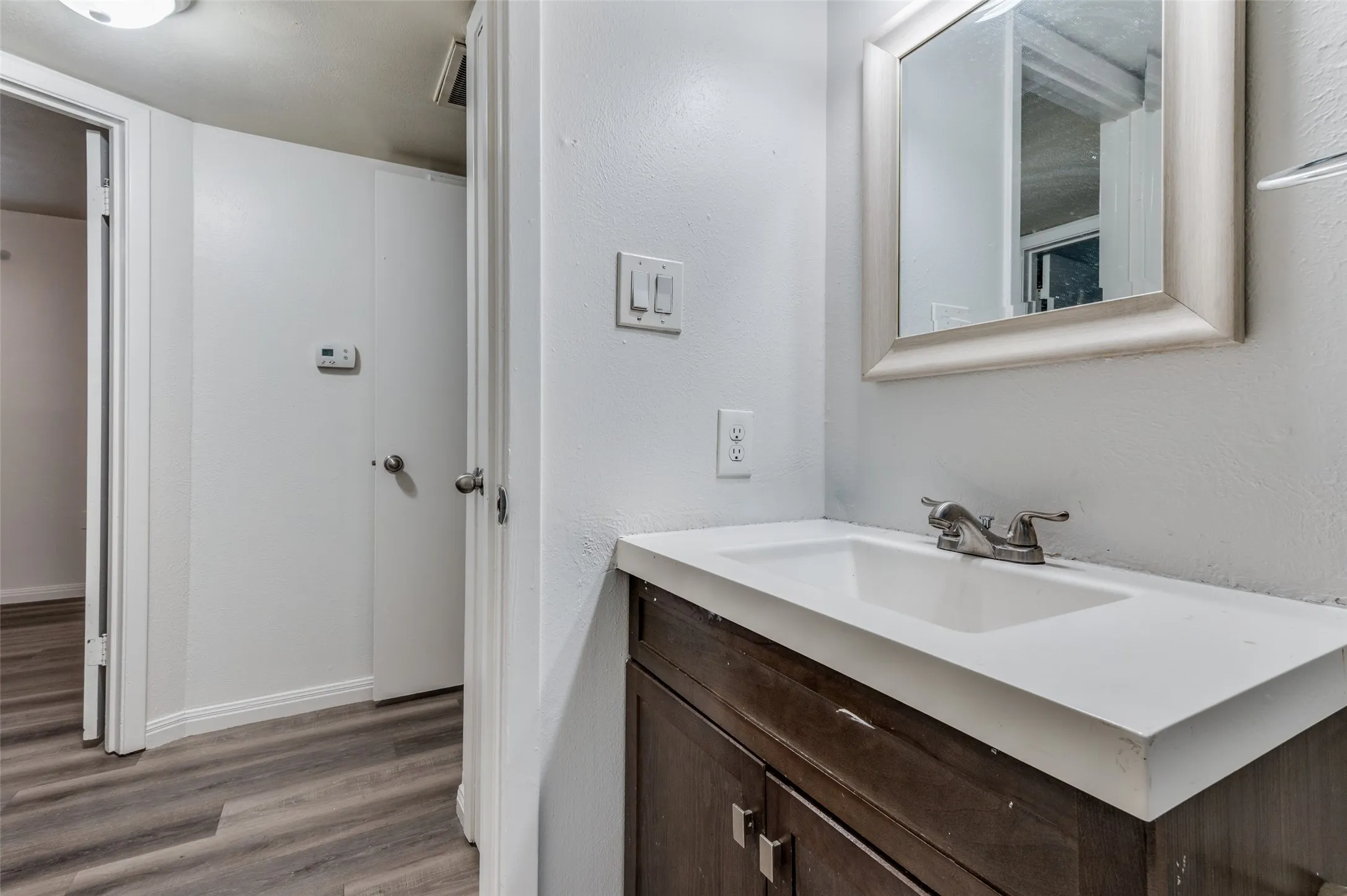 Wood-finish flooring, white walls, dark wood-finish vanity with an integrated sink, silver-finish faucet, and a framed mirror