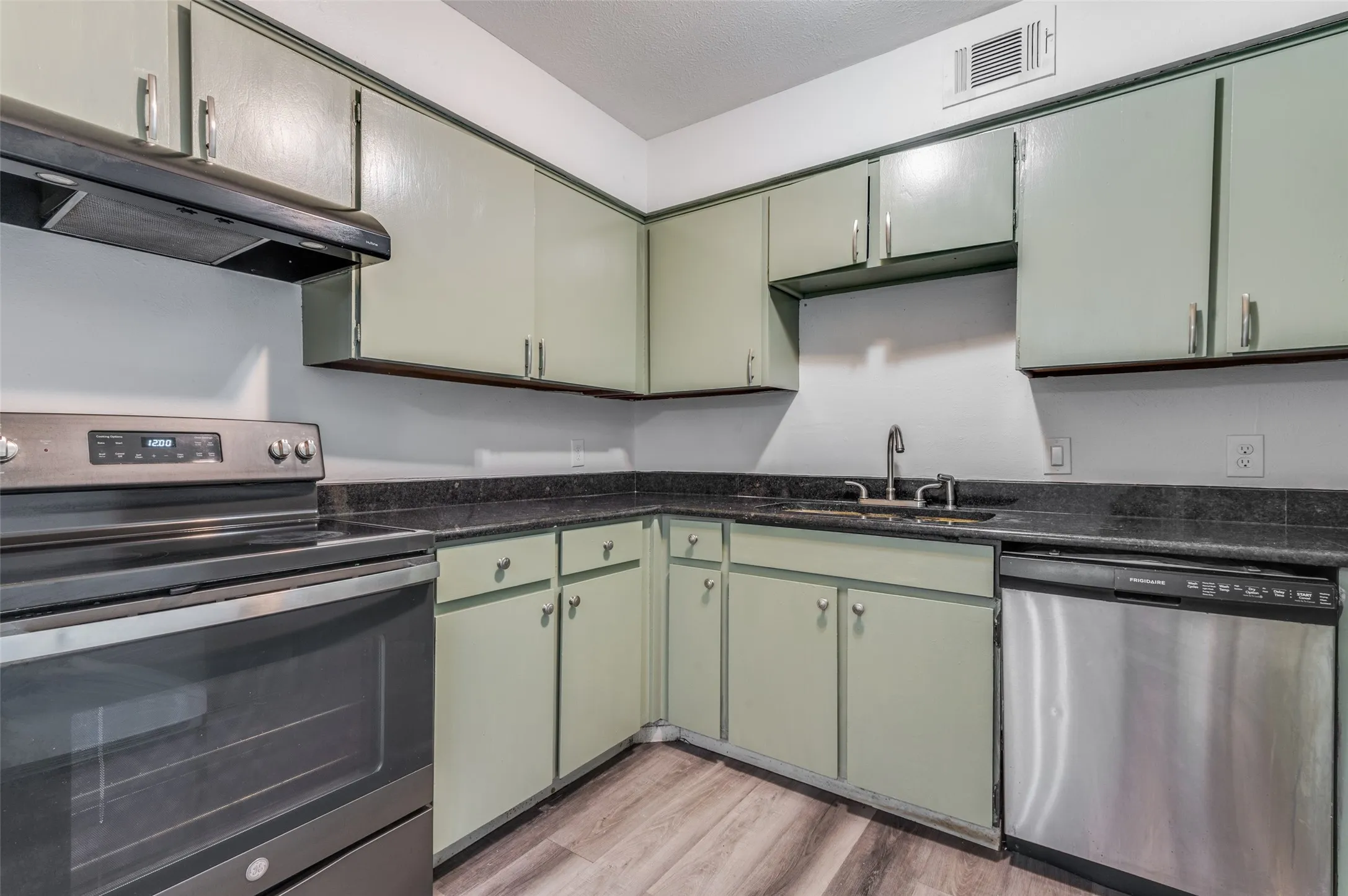 Kitchen featuring light green cabinetry, dark countertops, wood-finish flooring, a stainless steel dishwasher, and a stainless steel range