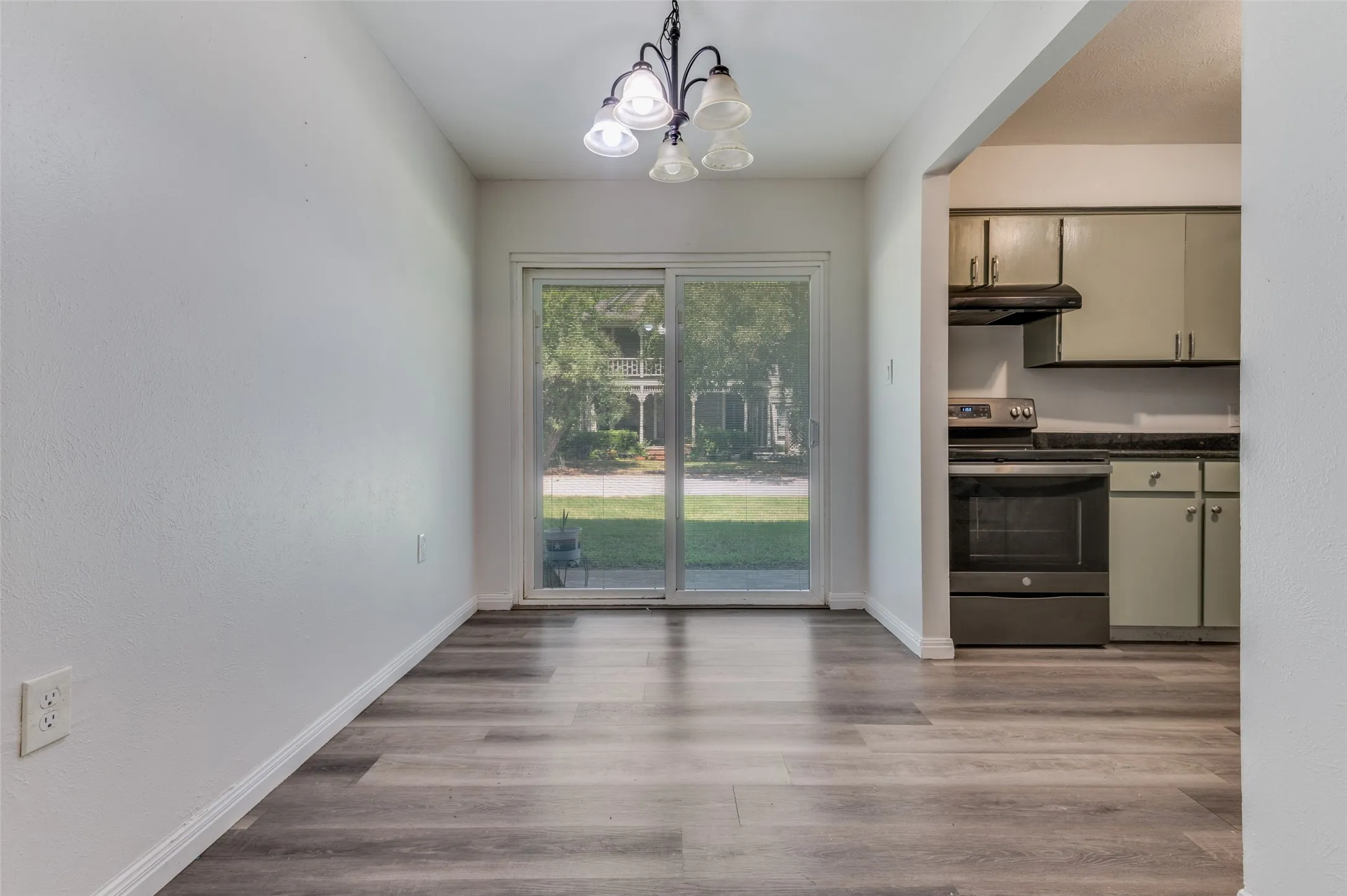 Open concept living space featuring wood-finish flooring, abundant natural light from glass sliding doors overlooking the landscaped grounds, a contemporary pendant light fixture, and an adjacent kitchen with stainless steel appliances and neutral cabinetry