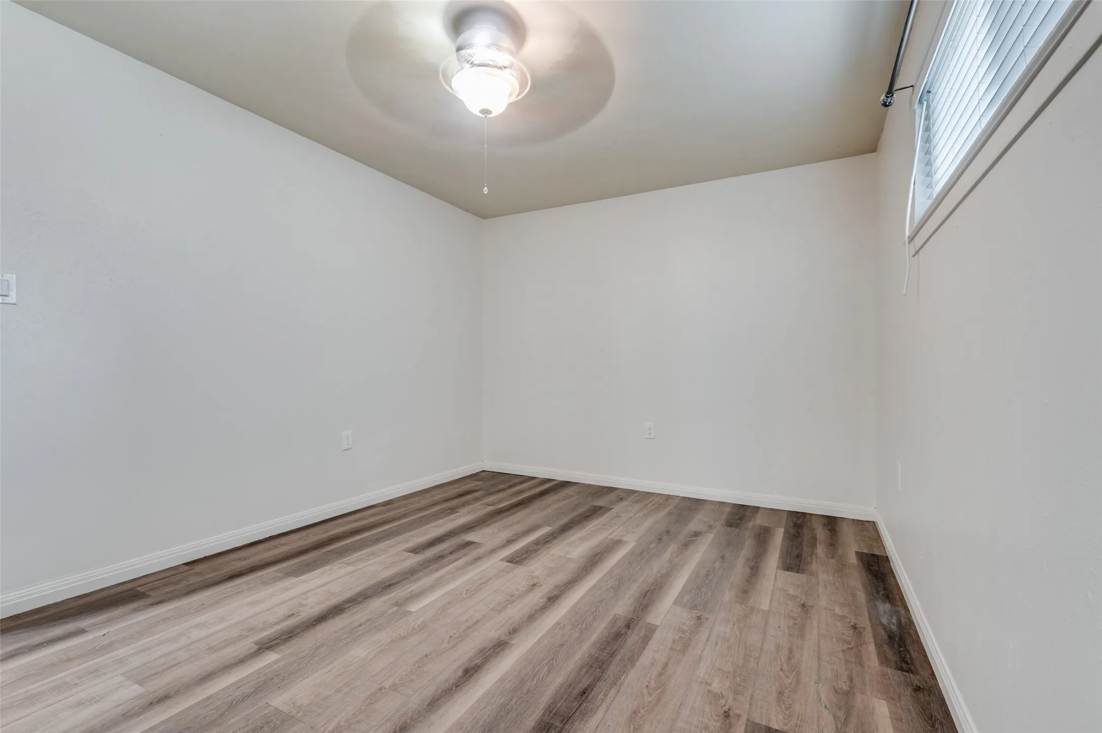 Wood-finish flooring, white walls, ceiling fan with integrated lighting, and window with blinds
