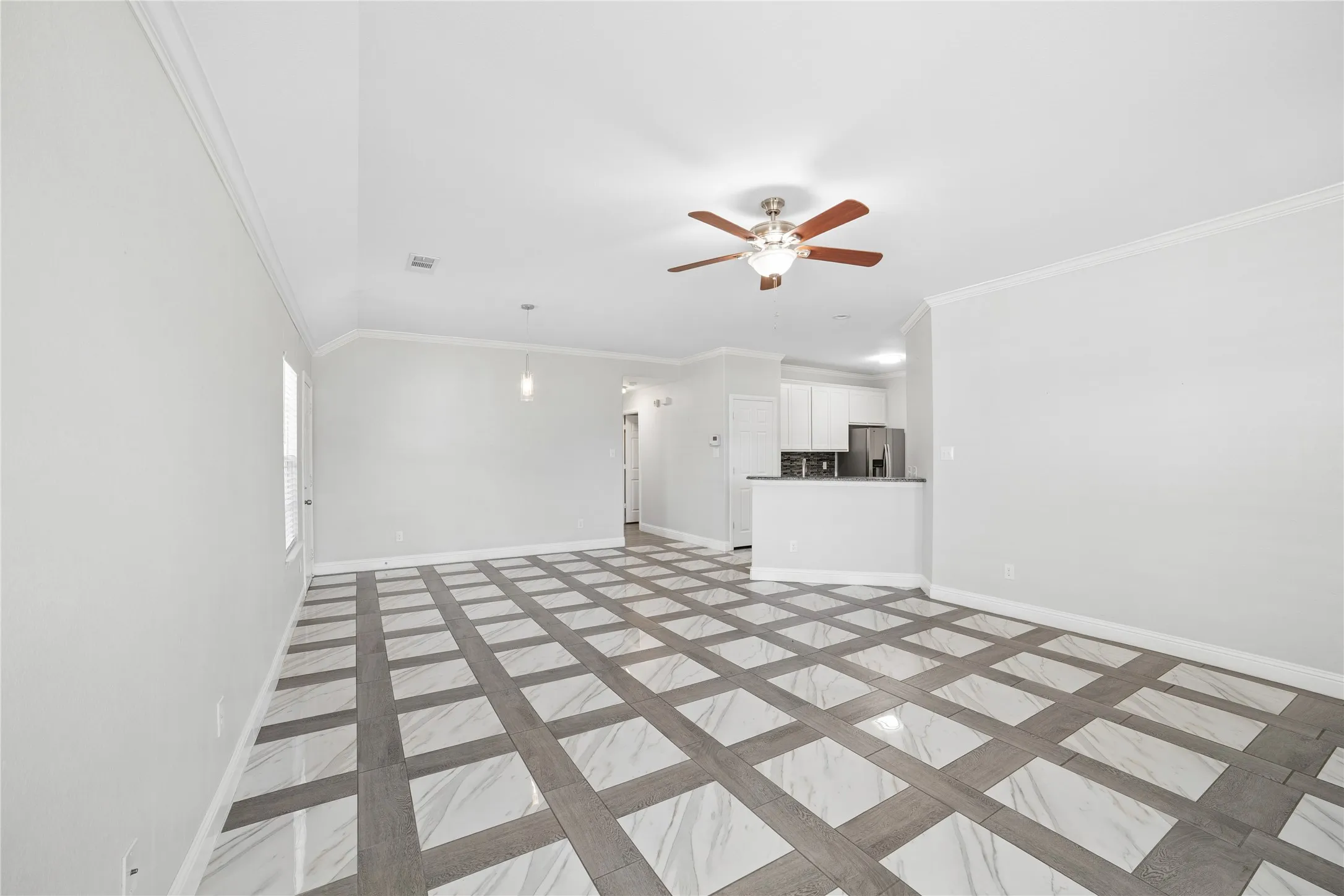 Spacious living area featuring patterned tile flooring, a ceiling fan with light, crown molding, and a kitchen pass-through with a breakfast bar