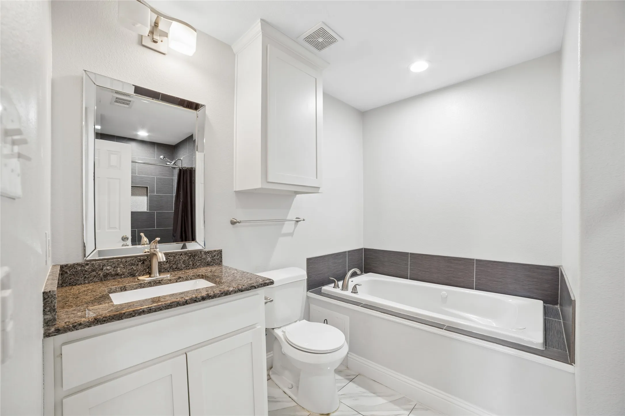 Modern bathroom featuring a granite-topped vanity with integrated sink, framed mirror with integrated lighting, white cabinetry, ceramic tile flooring, a soaking tub with dark tile surround and chrome fixtures, and a separate toilet compartment with wall-mounted storage
