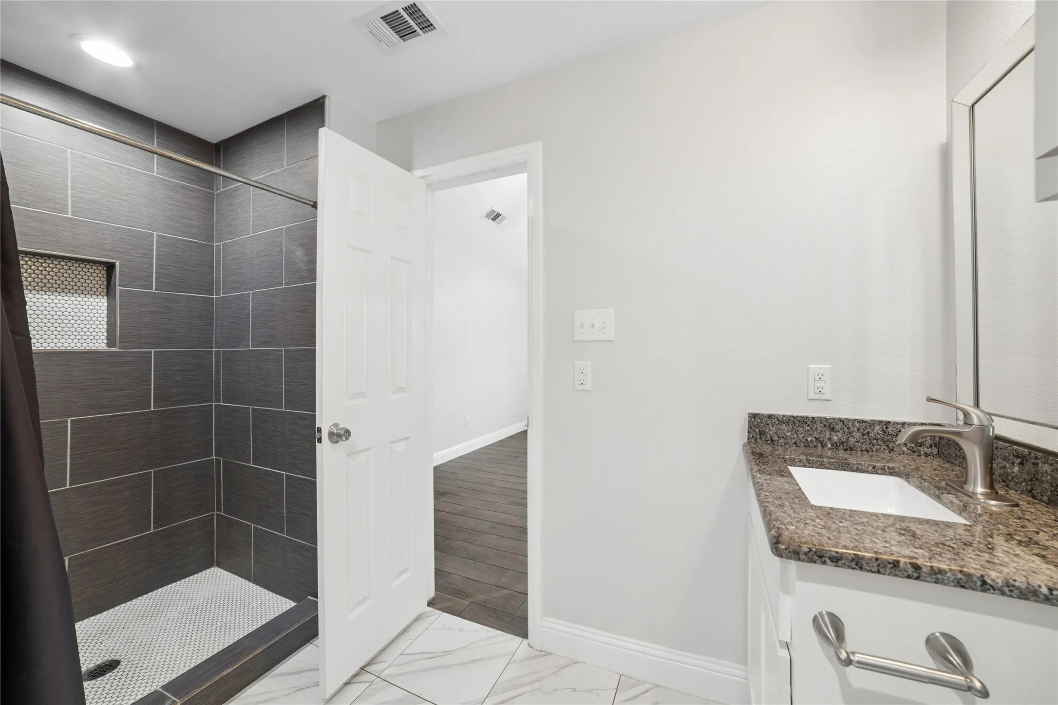 Shower with dark gray tile surround and integrated niche, white penny tile shower floor, vanity with granite countertop and undermount sink, and brushed nickel faucet
