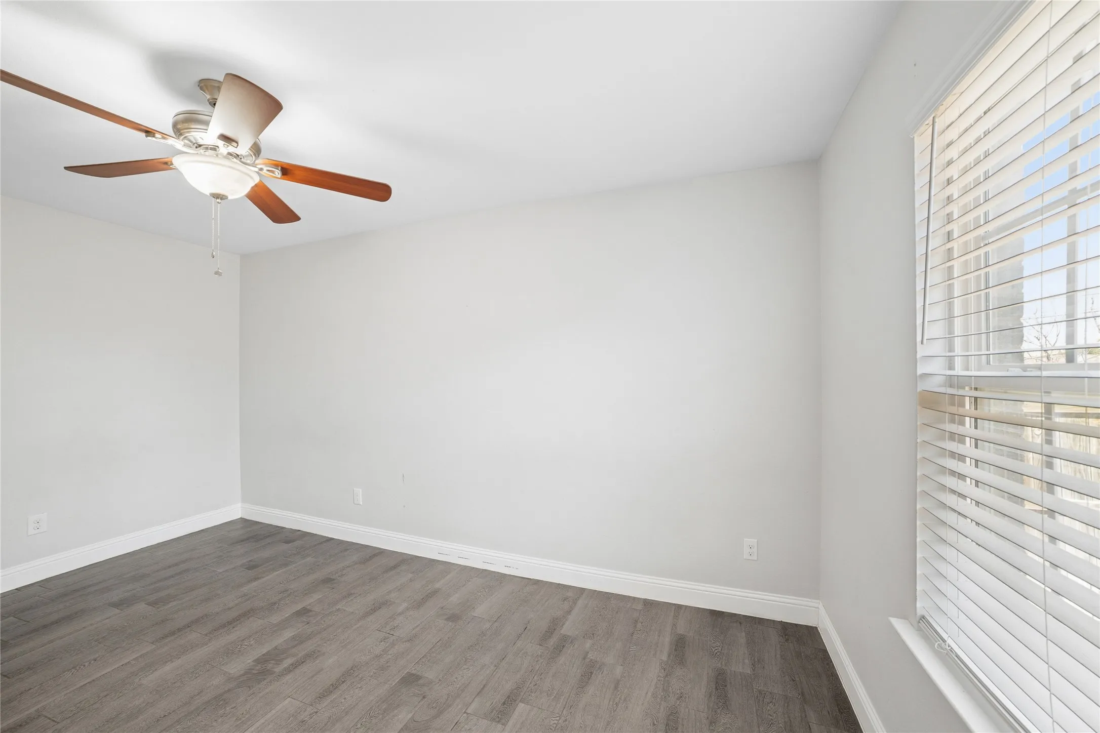 Wood-finish flooring, white walls, ceiling fan with integrated lighting, and window with blinds