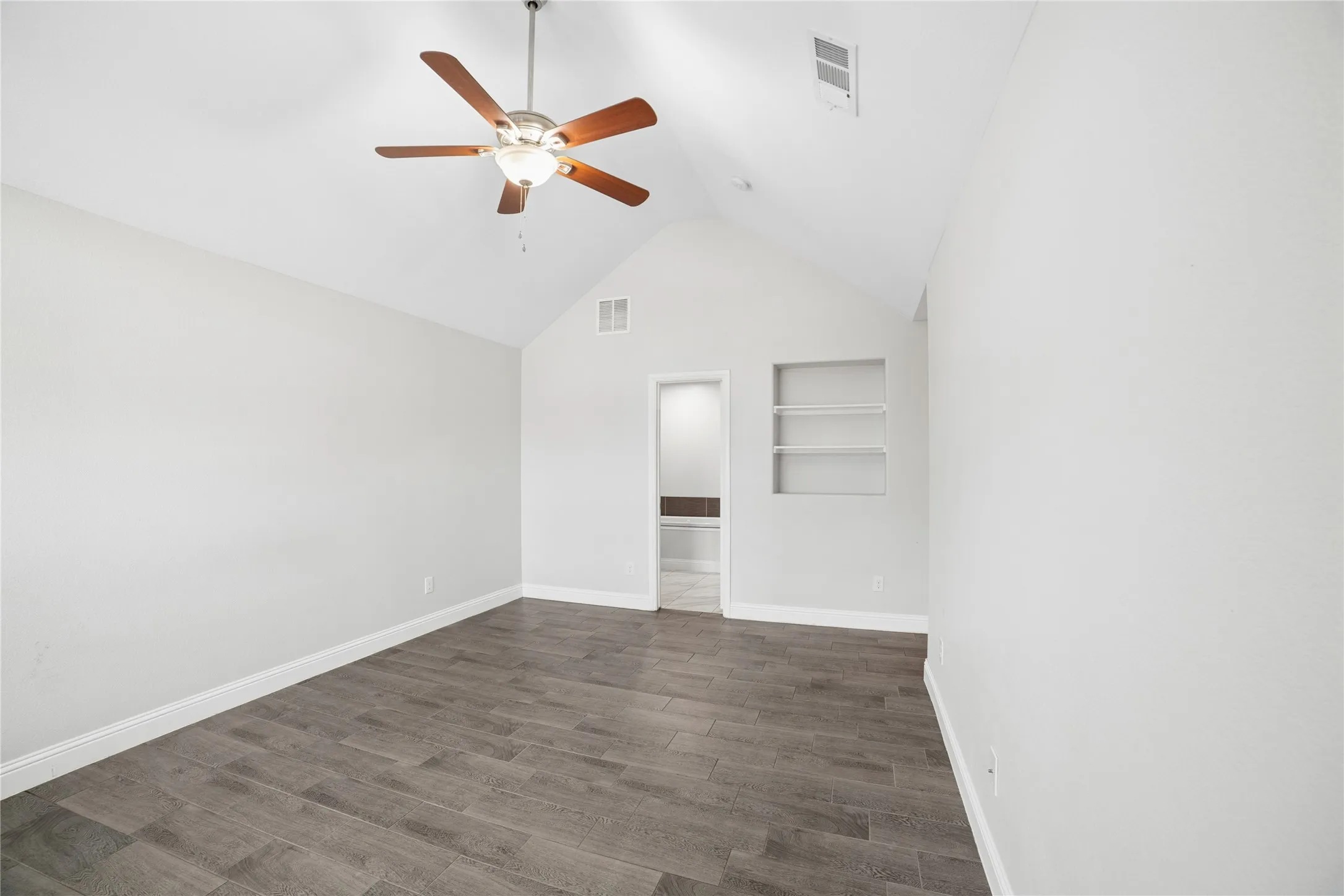 Vaulted ceiling with a ceiling fan, wood-finish flooring, recessed shelving, and a doorway opening