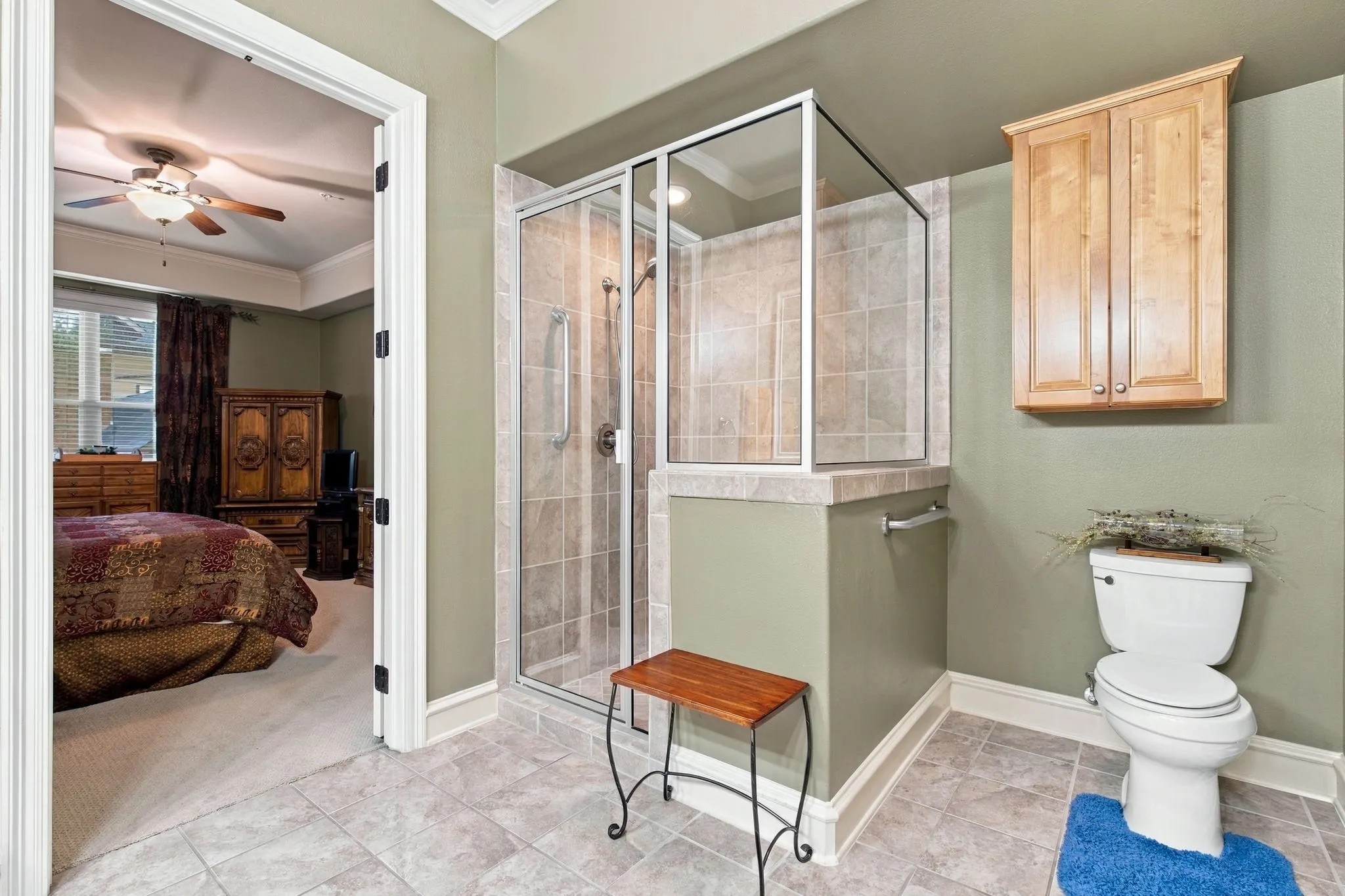Tiled flooring, glass-enclosed shower with tiled surround, crown molding, toilet with wood-finish wall-mounted cabinetry
