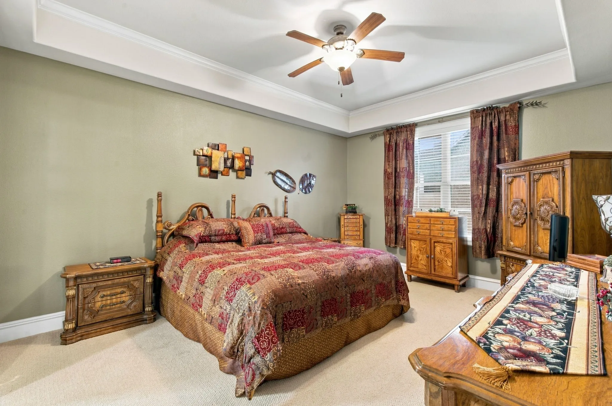Tray ceiling with crown molding, ceiling fan with integrated lighting, window with blinds, and neutral carpeting