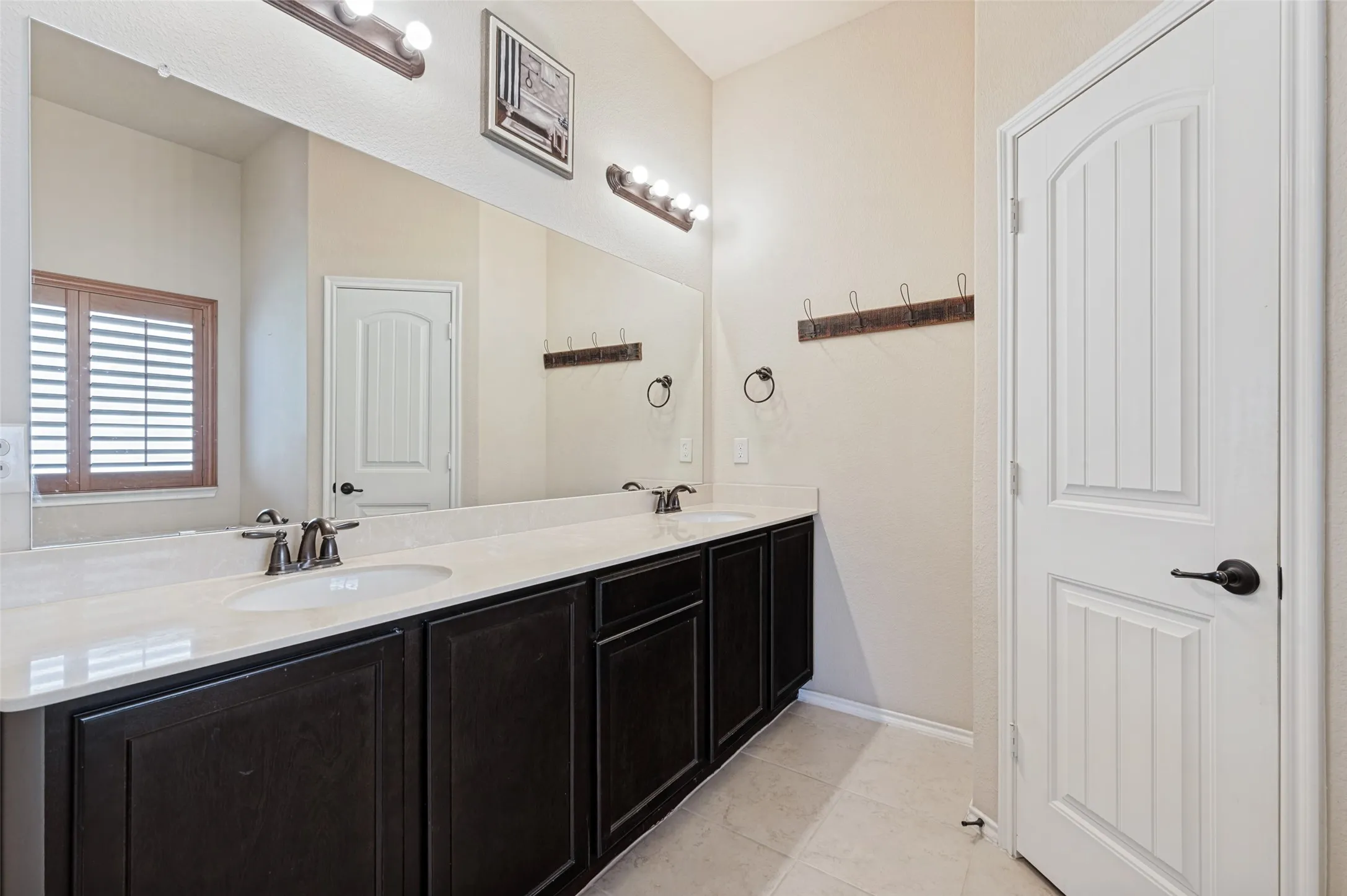 Dual undermount sinks with brushed bronze fixtures, dark wood-finish cabinetry, light-toned countertop, large wall-mounted mirror, and wood plantation shutters