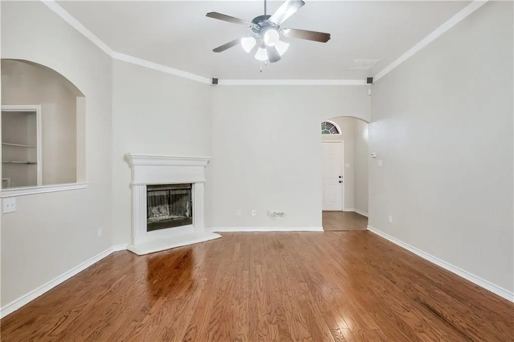 Spacious living room showcasing gleaming wood-finish flooring, a decorative fireplace with white surround and hearth, ceiling fan with integrated lighting, and abundant natural light from multiple windows