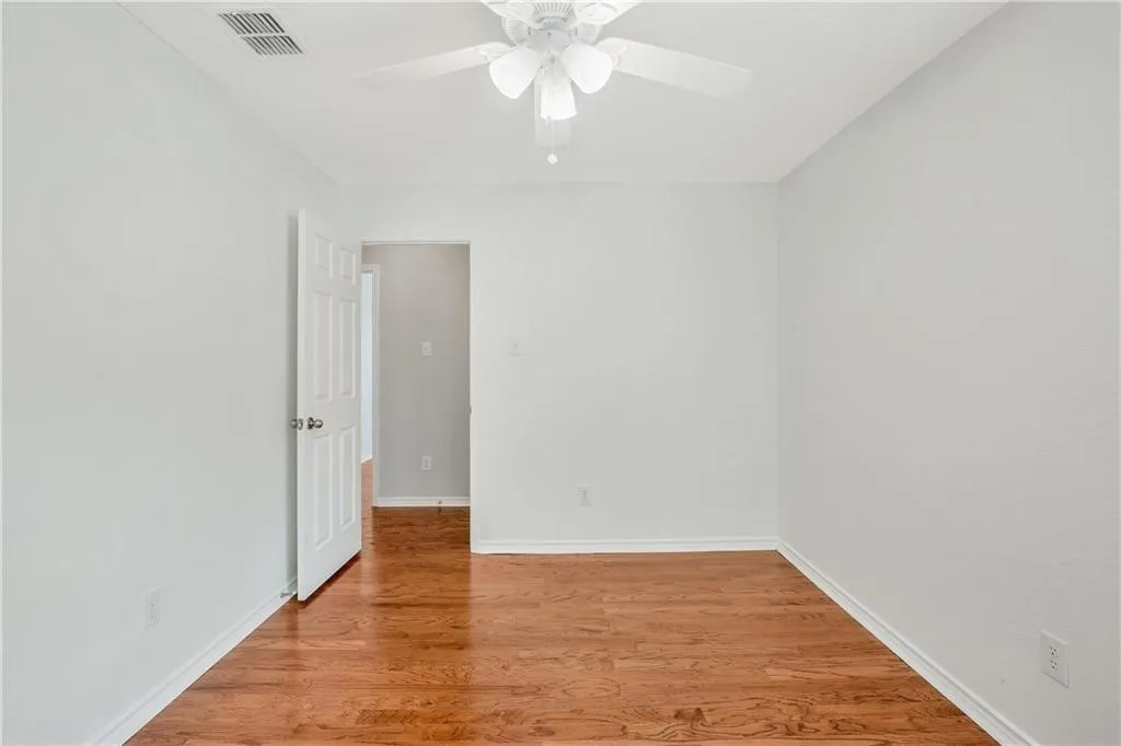 Spacious bedroom with warm wood-finish flooring, white painted walls, ceiling fan with integrated lighting, and direct access to an adjoining room through a white six-panel door