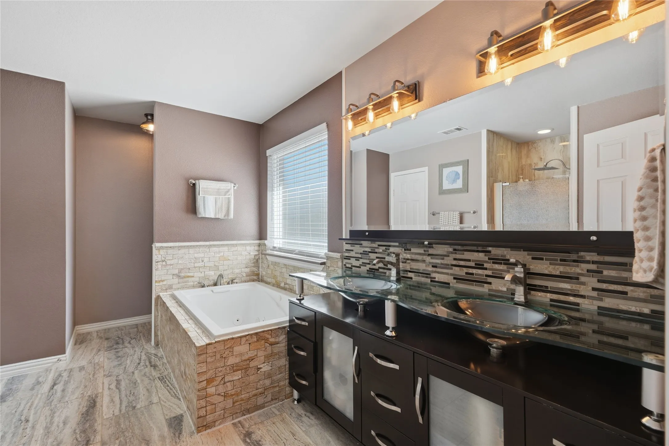 Dual vessel sinks, stone tile backsplash, dark wood-finish vanity, built-in jetted tub, and large format tile flooring